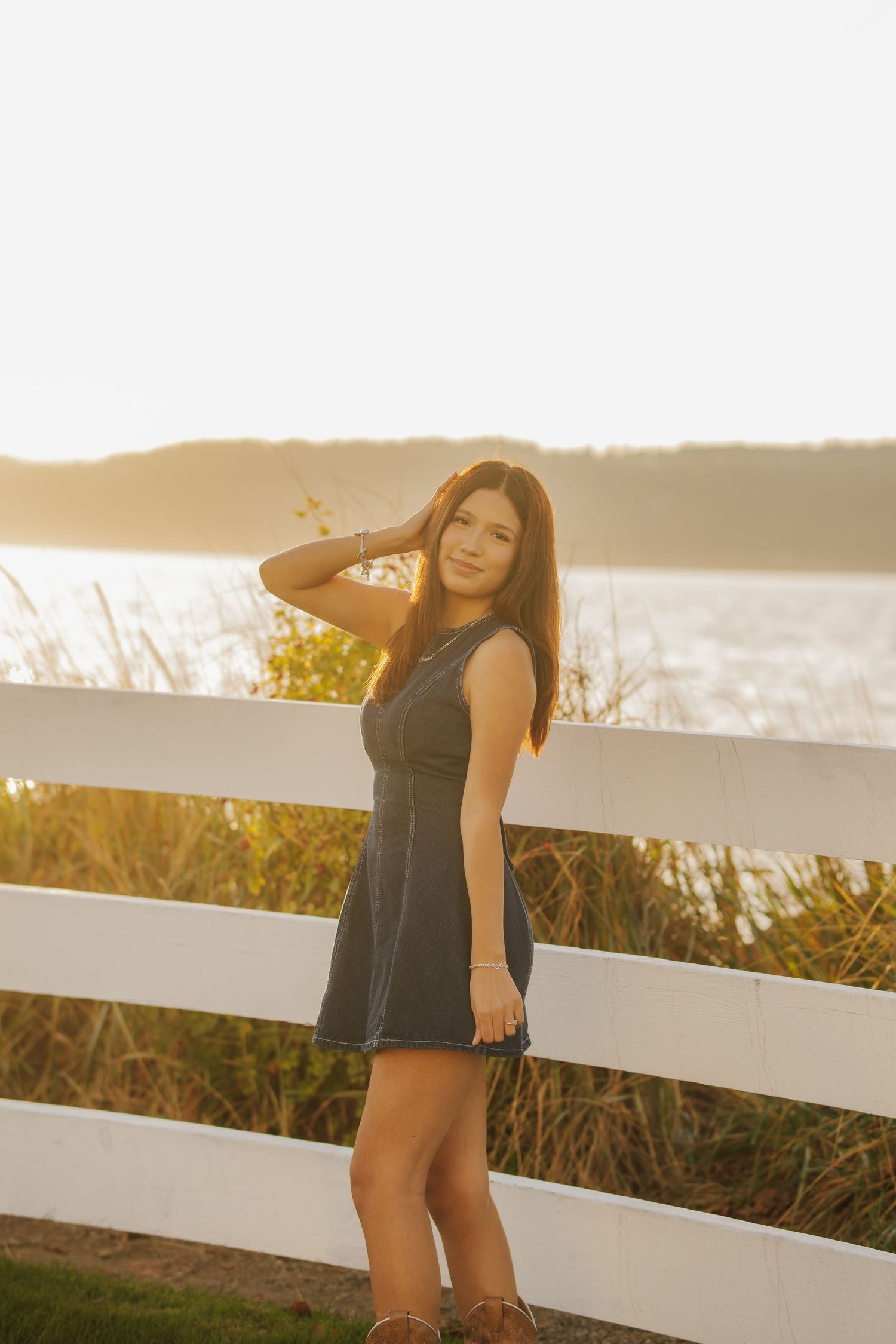 Young woman in a gray sleeveless dress poses with her hand in her hair beside a white wooden fence overlooking water at golden hour.