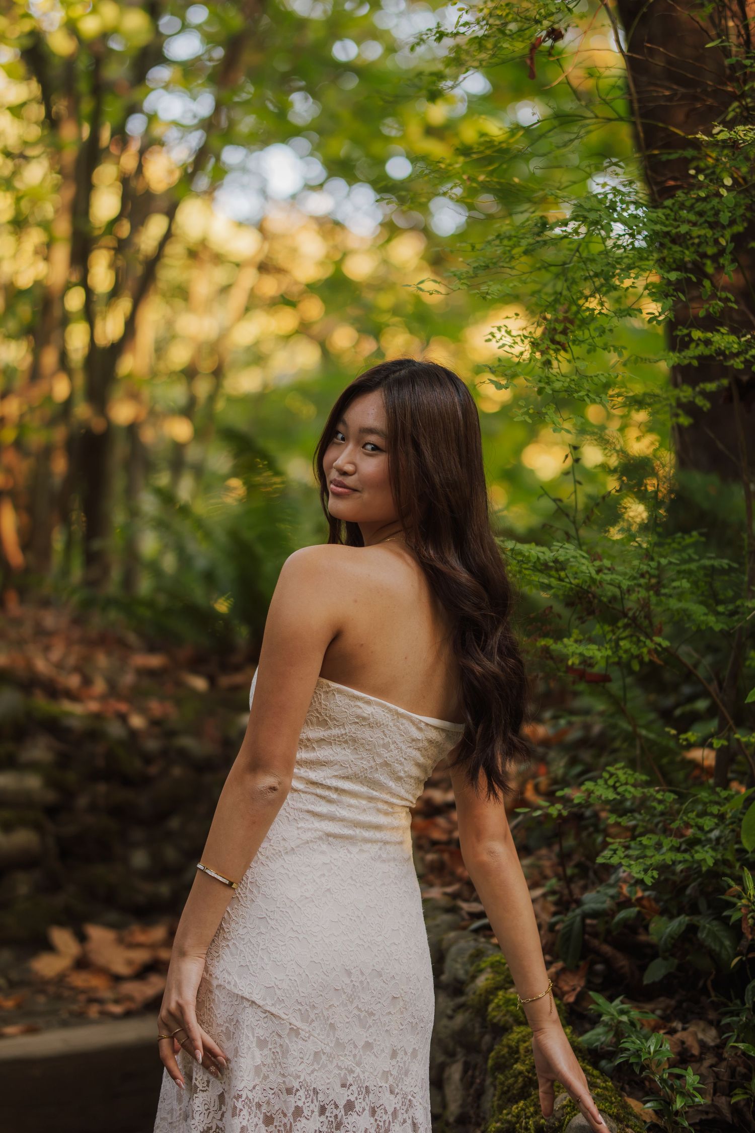 Woman with long dark hair in a white lace strapless dress looks over her shoulder in a sunlit forest with autumn leaves.