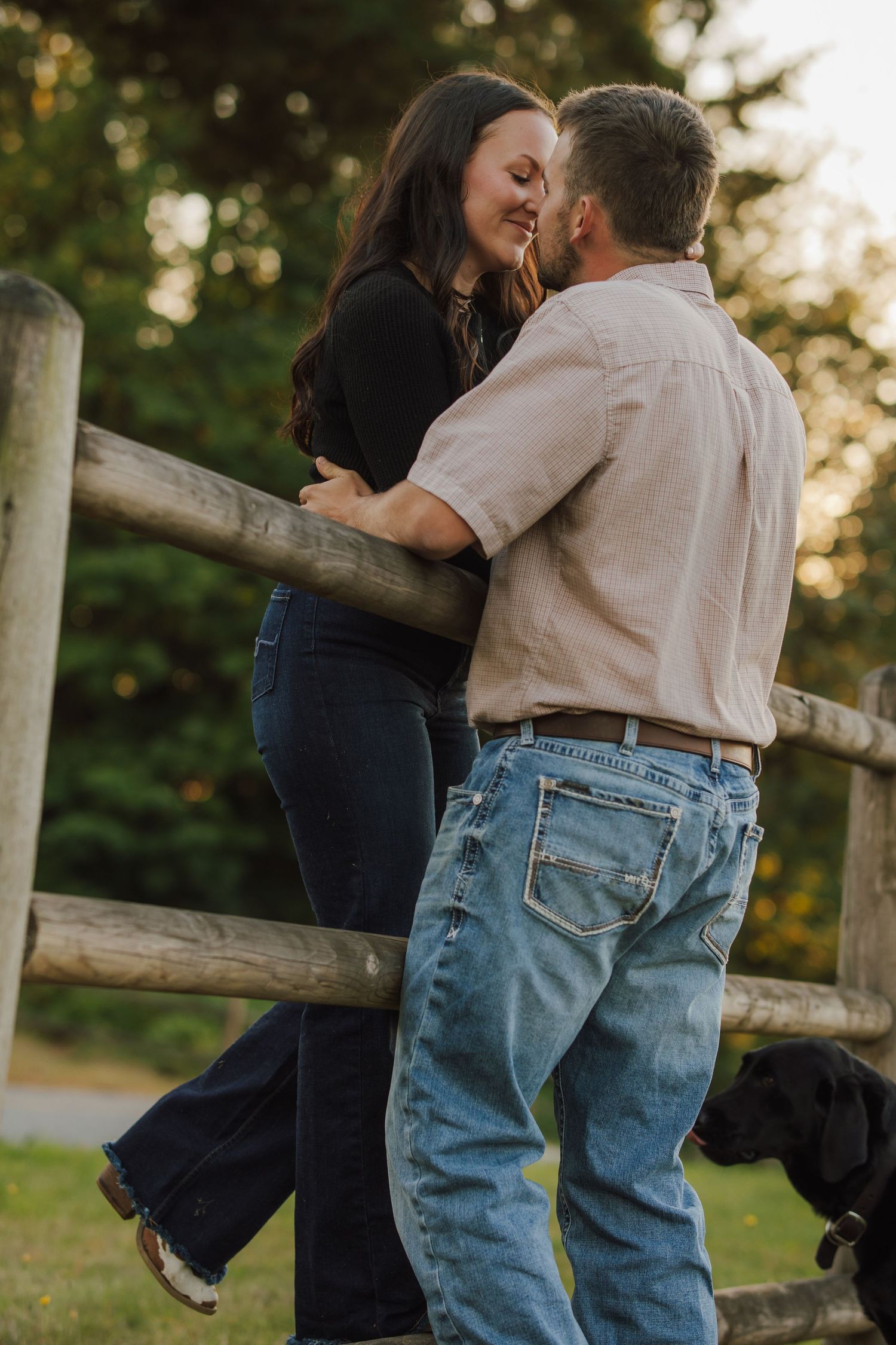 Couple in denim embracing and smiling beside a rustic wooden fence at golden hour, with a black Labrador resting nearby.