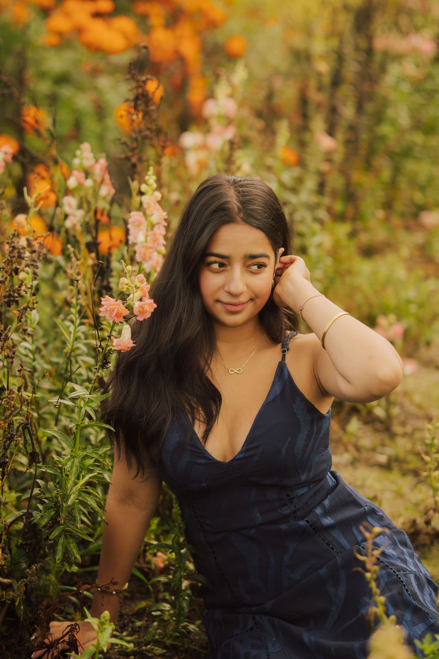 Young woman with long dark hair in a navy dress sits among blooming orange and pink flowers in a sunlit garden, touching her hair.