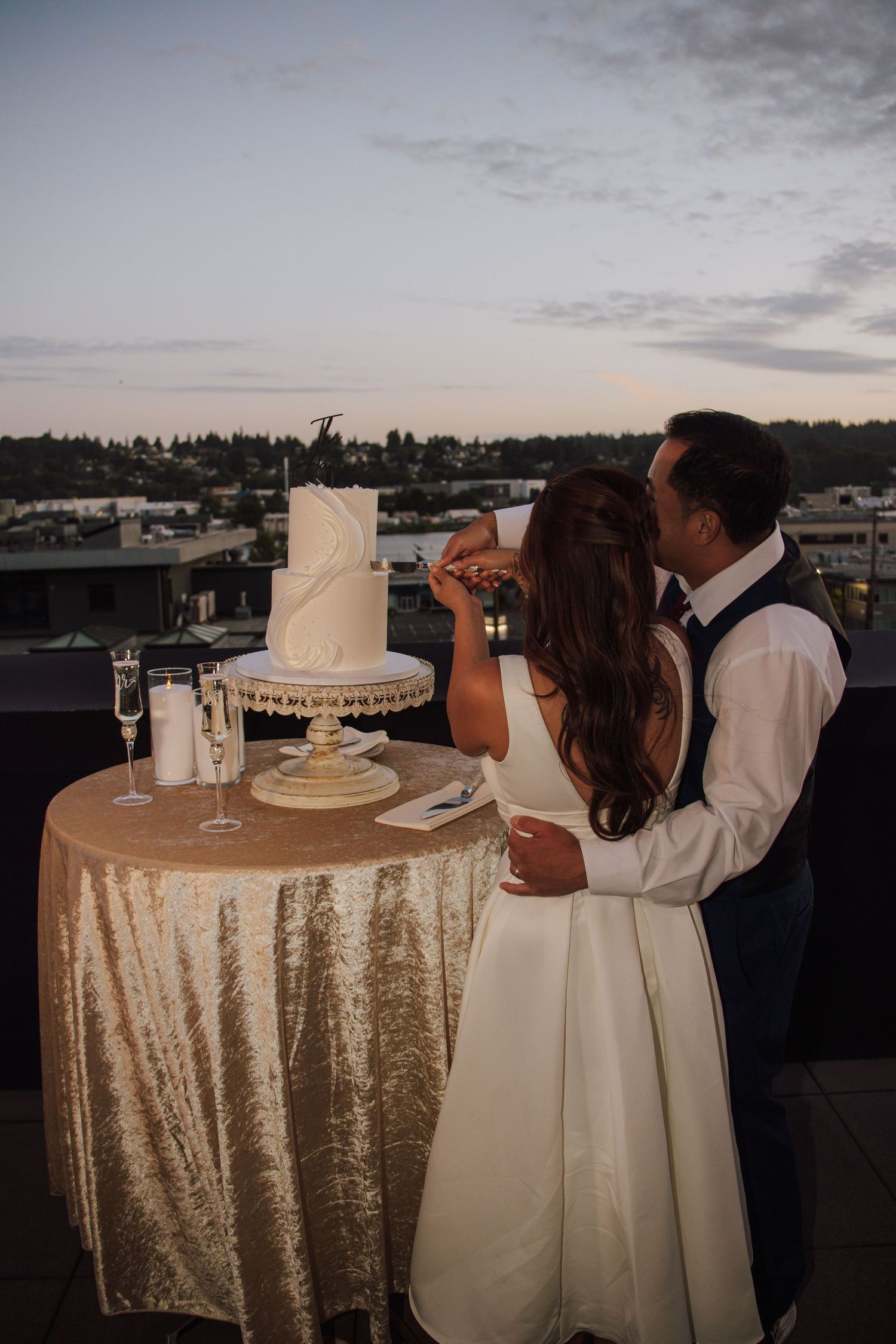 Couple cuts tiered white wedding cake on rooftop at dusk with city skyline backdrop.