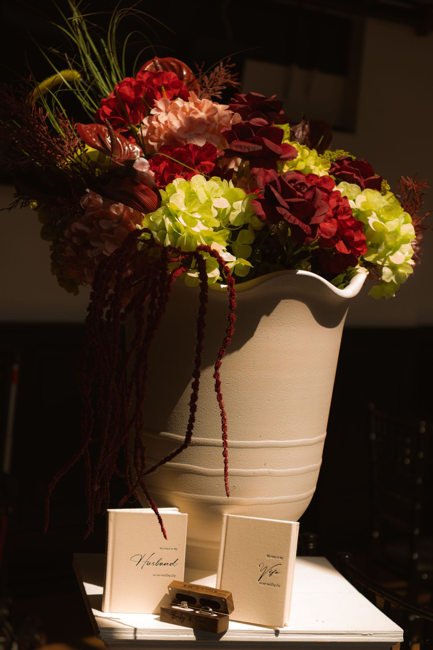 Lush floral arrangement of red roses and green hydrangeas in white vase with wedding stationery.