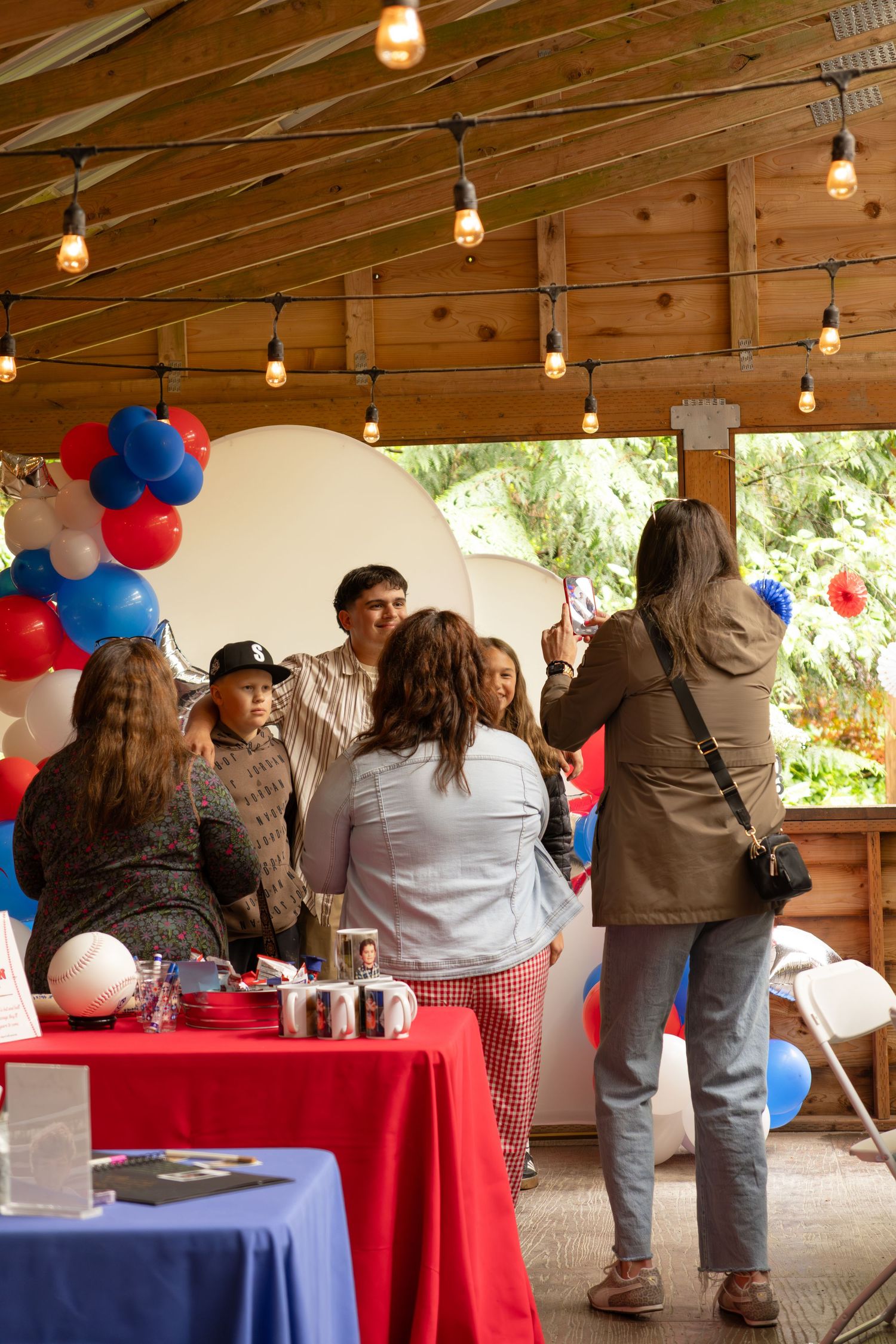 Party guests posing for photos near red, white, and blue balloon arch at celebration event.