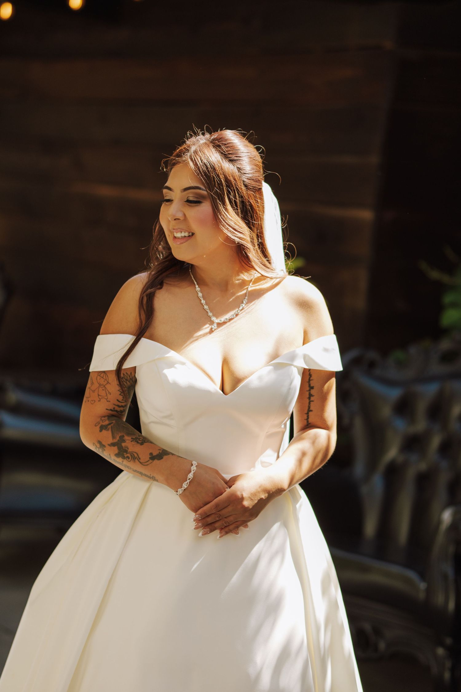 Radiant bride in off-shoulder white satin wedding gown with tattoos and pearl necklace in sunlight.
