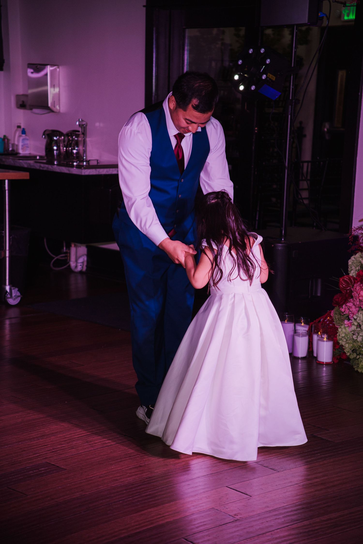 Groom dances with young flower girl in white dress on wedding reception dance floor.
