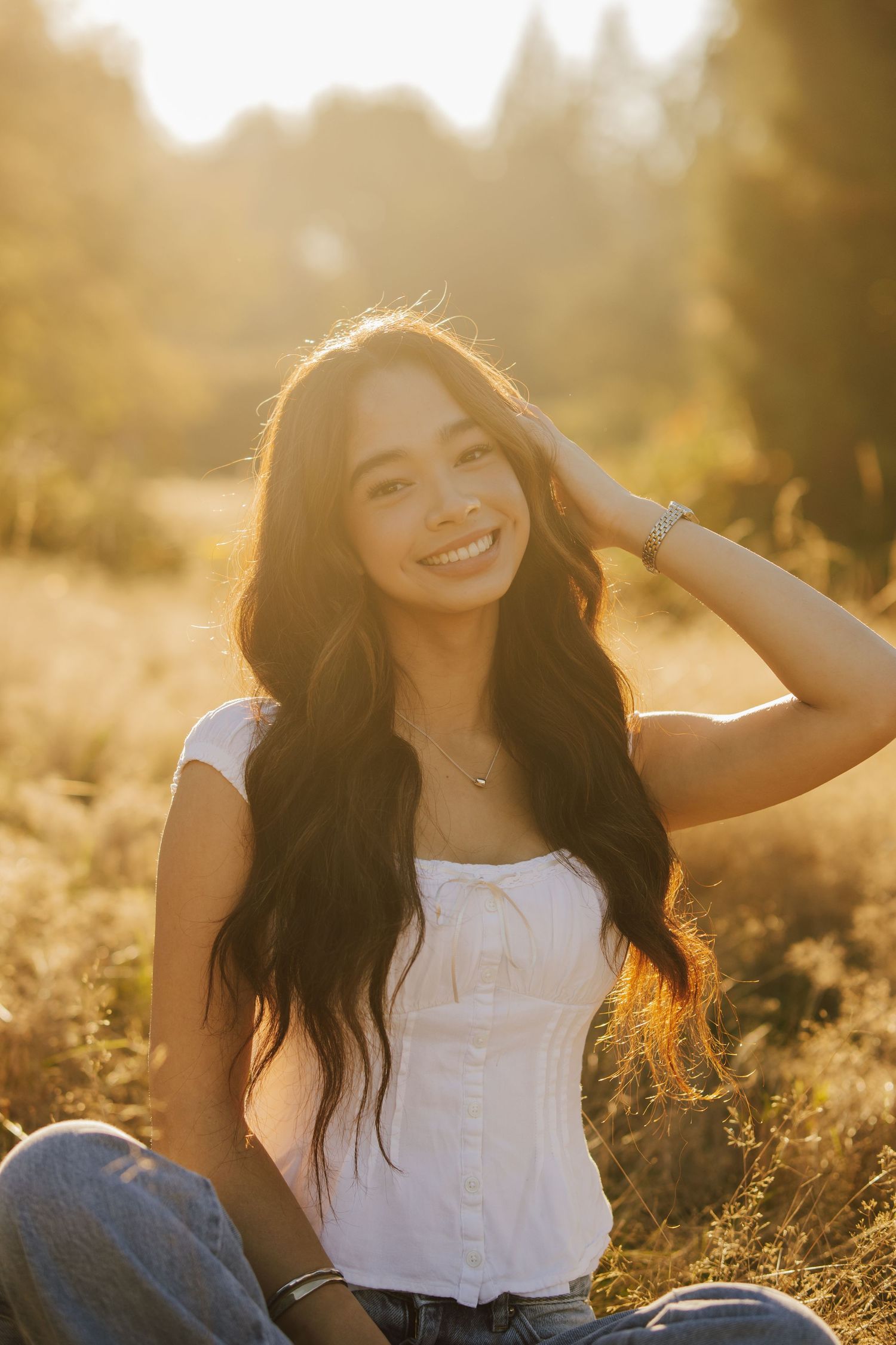 Smiling young woman with long wavy dark hair in a white top sits in a golden sunlit field, hand raised to her head.
