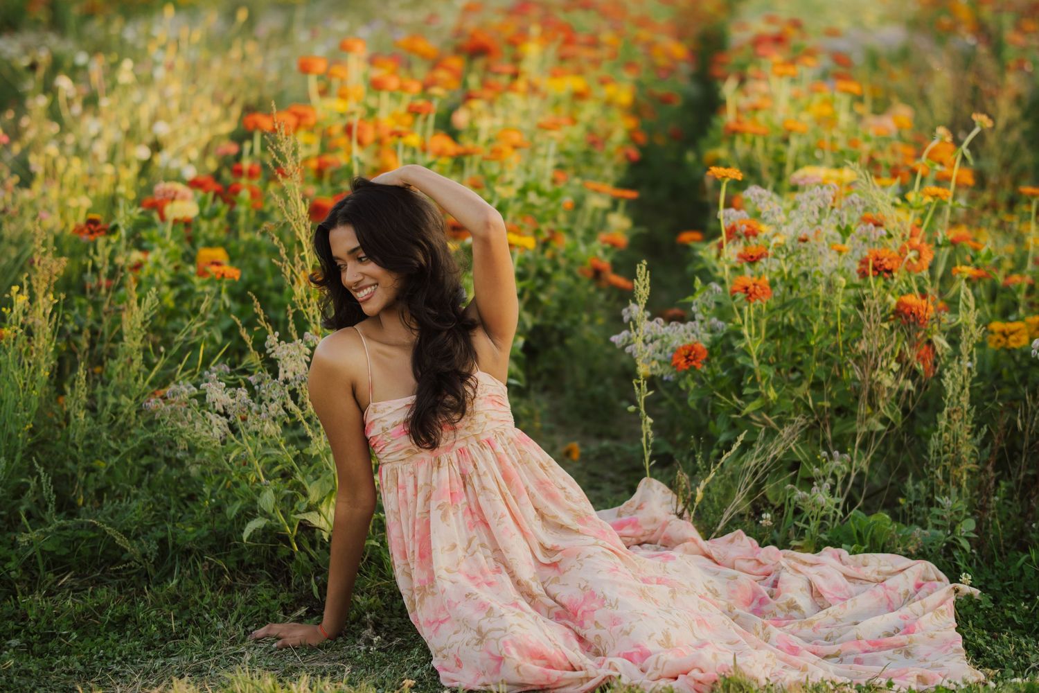 Woman with long dark hair in a flowing pink floral dress sitting on the grass among orange wildflowers in a sunlit meadow.