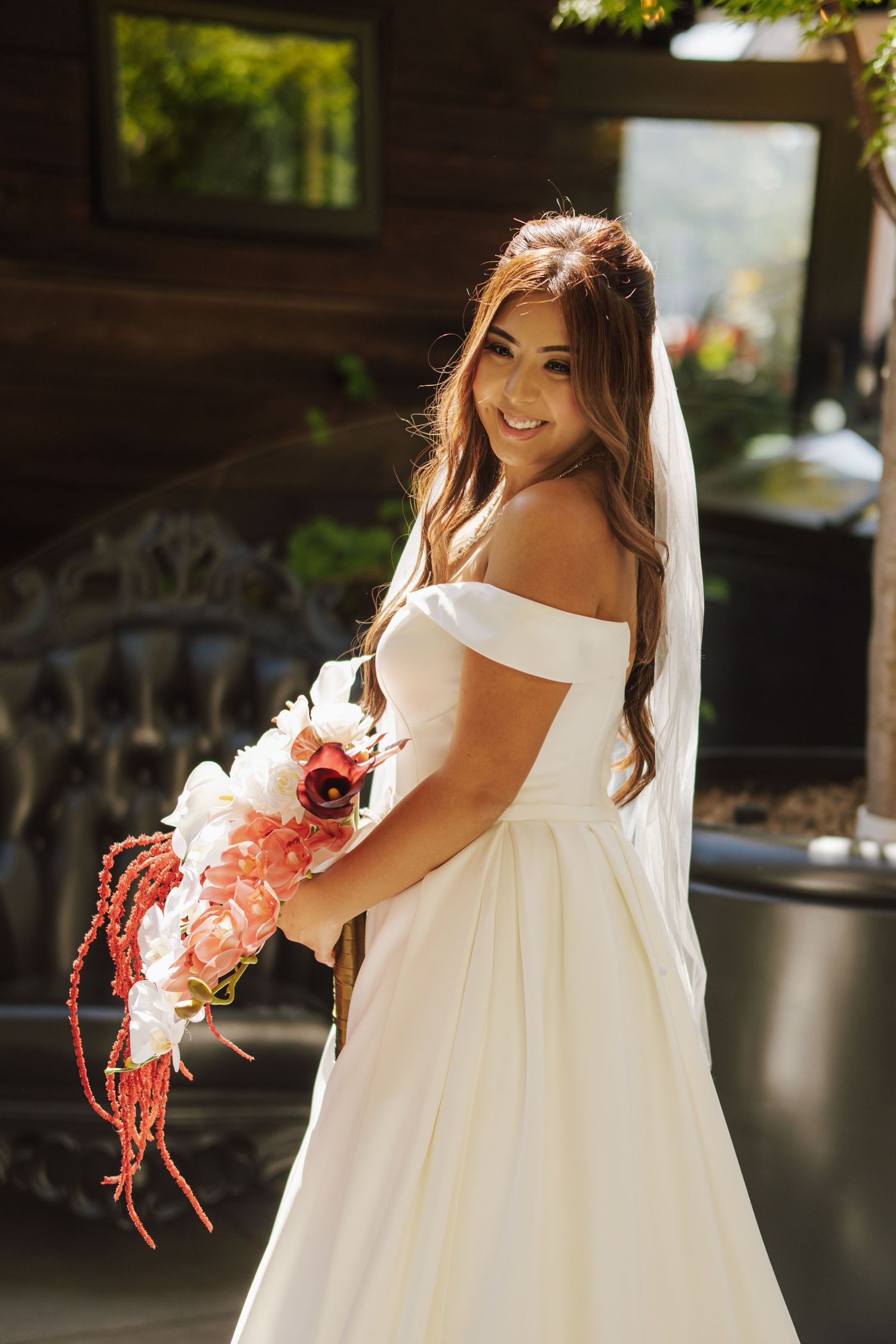 Smiling bride in off-shoulder white dress holding a pink and red floral bouquet.