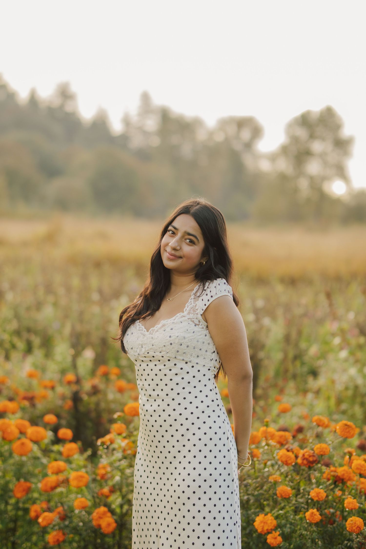 Woman in a white polka dot dress smiling in a field of orange marigold flowers at golden hour with trees in the background.
