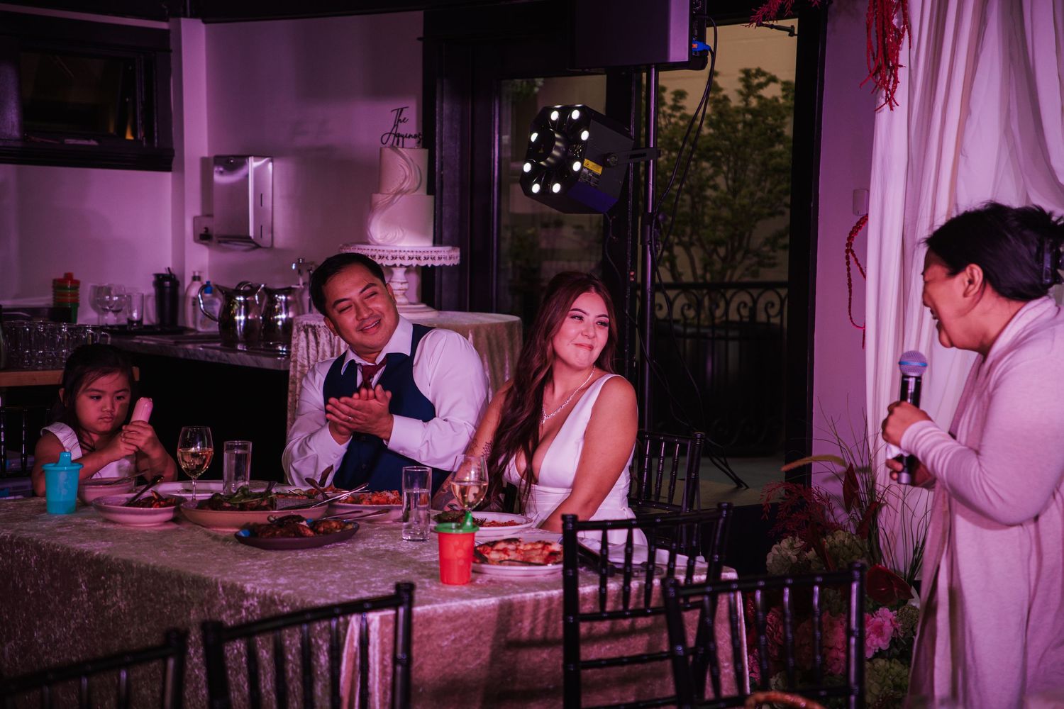 Bride and groom smile at dinner table during wedding reception speech under purple lighting.