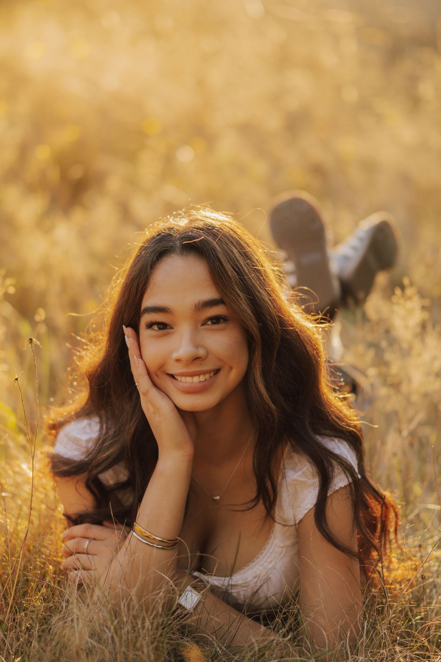 Smiling young woman with long wavy hair lies on her stomach in a golden sunlit meadow, resting her chin on her hand at sunset.