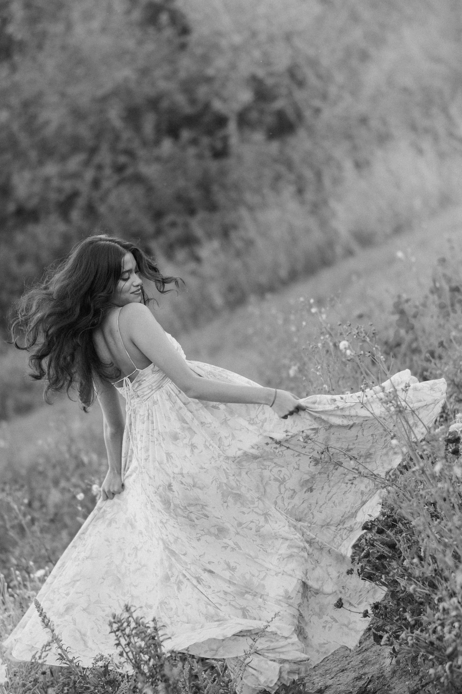 Black and white photo of a woman with long wavy hair twirling in a flowing floral dress in a wildflower meadow.