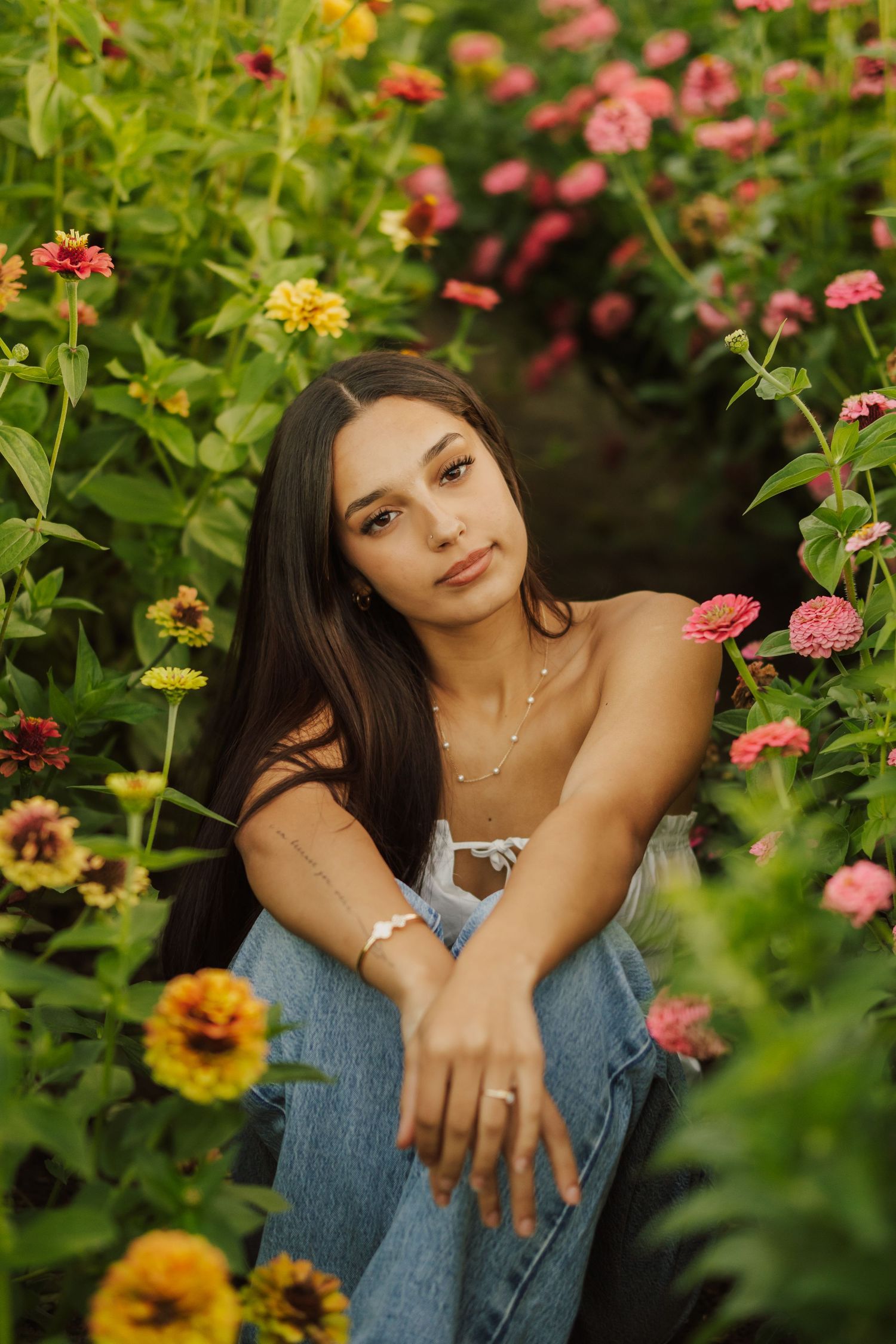 Young woman with long dark hair sits among blooming pink, yellow and orange zinnias in a sunlit flower garden, wearing jeans and a necklace.