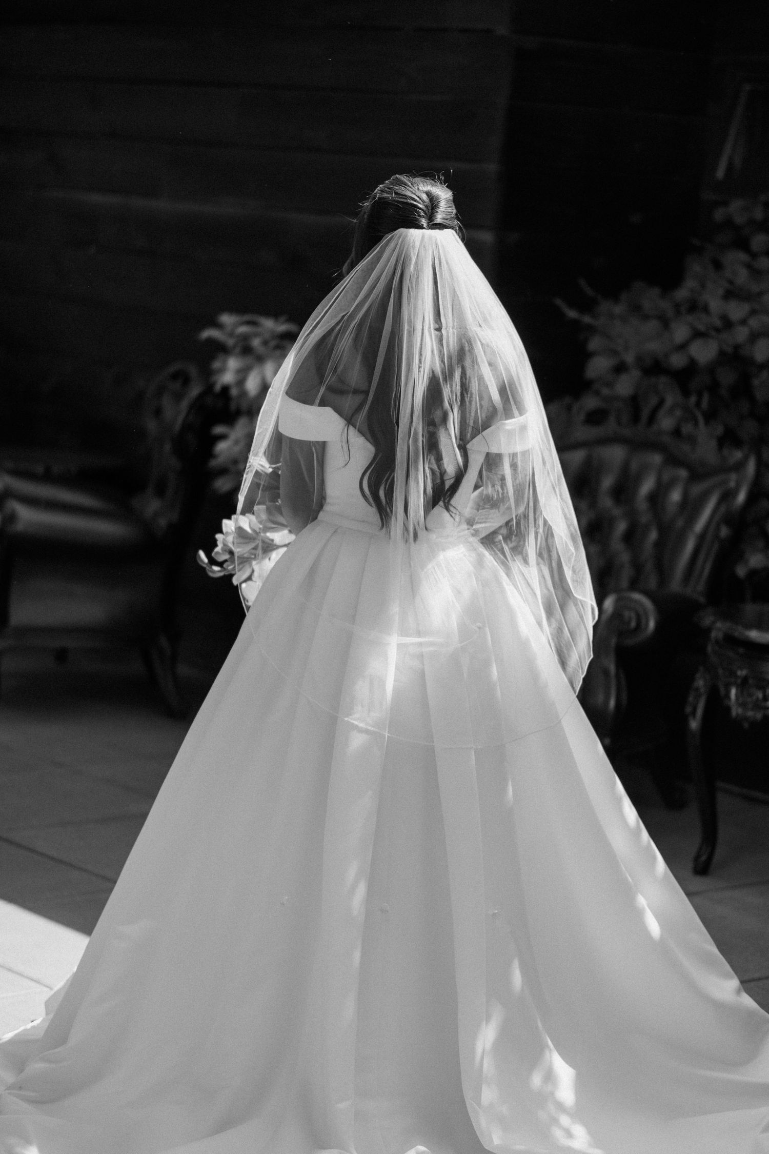 Black and white photo of a bride in a flowing white gown and veil seen from behind.