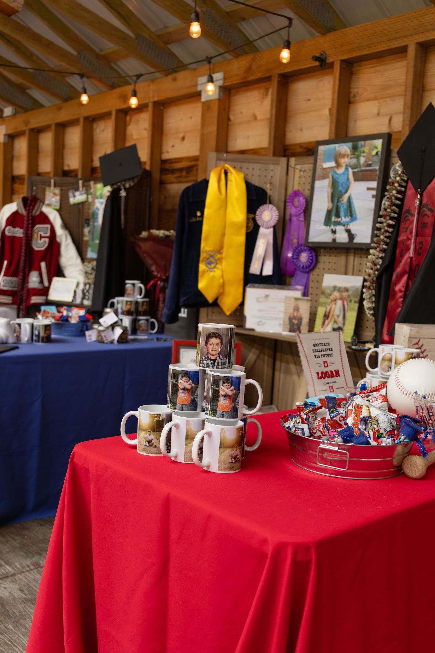 Graduation memorabilia display with personalized mugs, ribbons, and clothing on red and blue draped tables in a rustic barn setting.