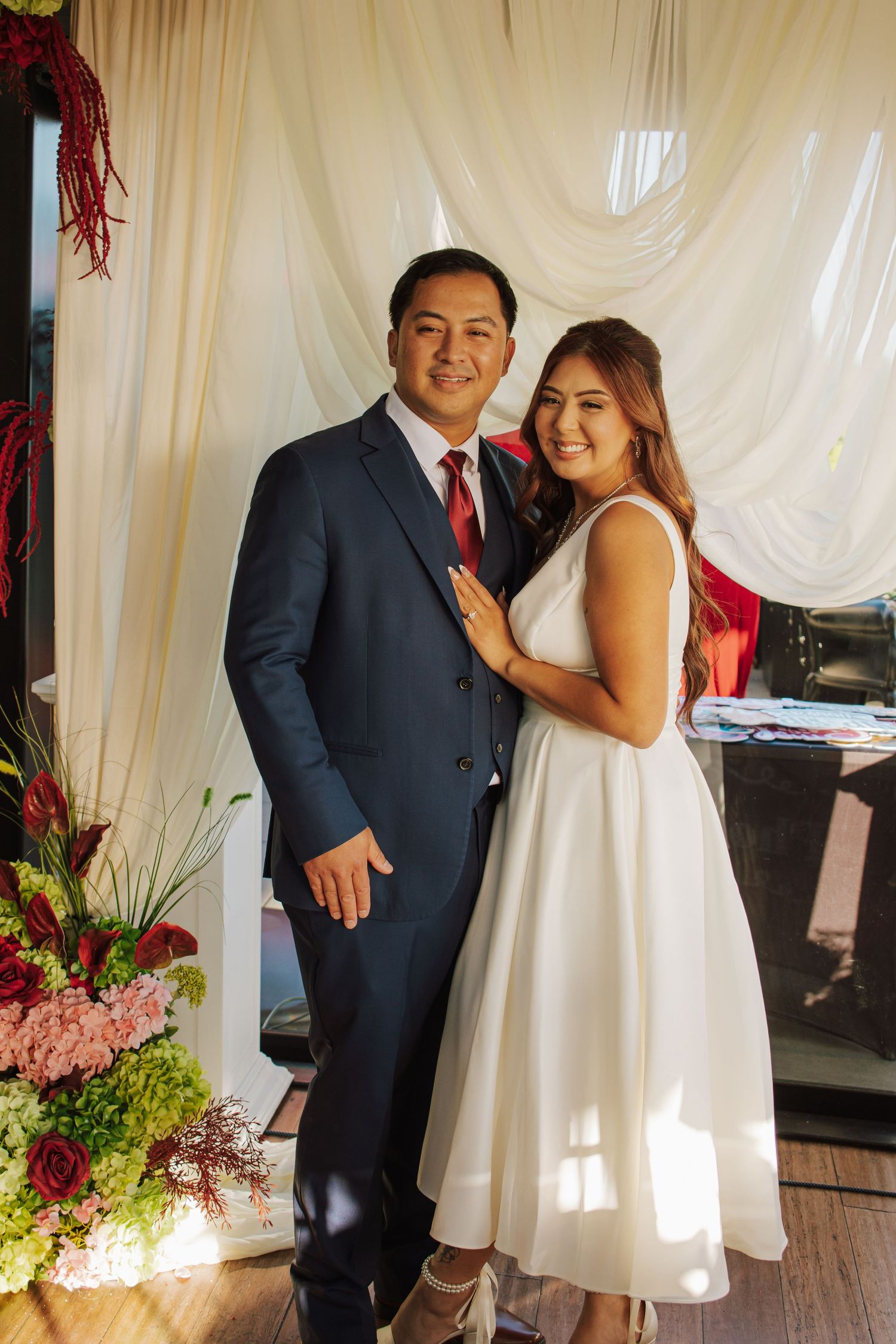 Bride in white midi dress and groom in navy suit pose together in front of floral backdrop.