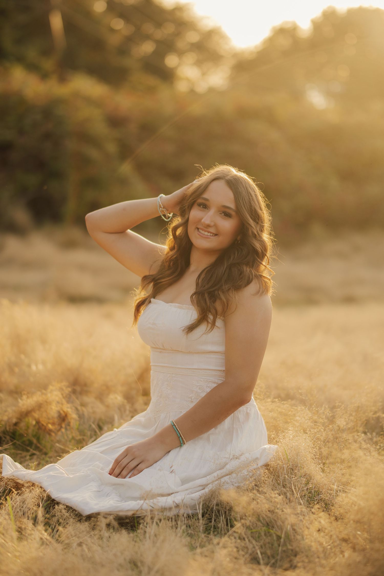 Smiling young woman in a white sundress sits in tall golden grass at sunset, one hand in her long wavy hair, with warm glowing light behind.