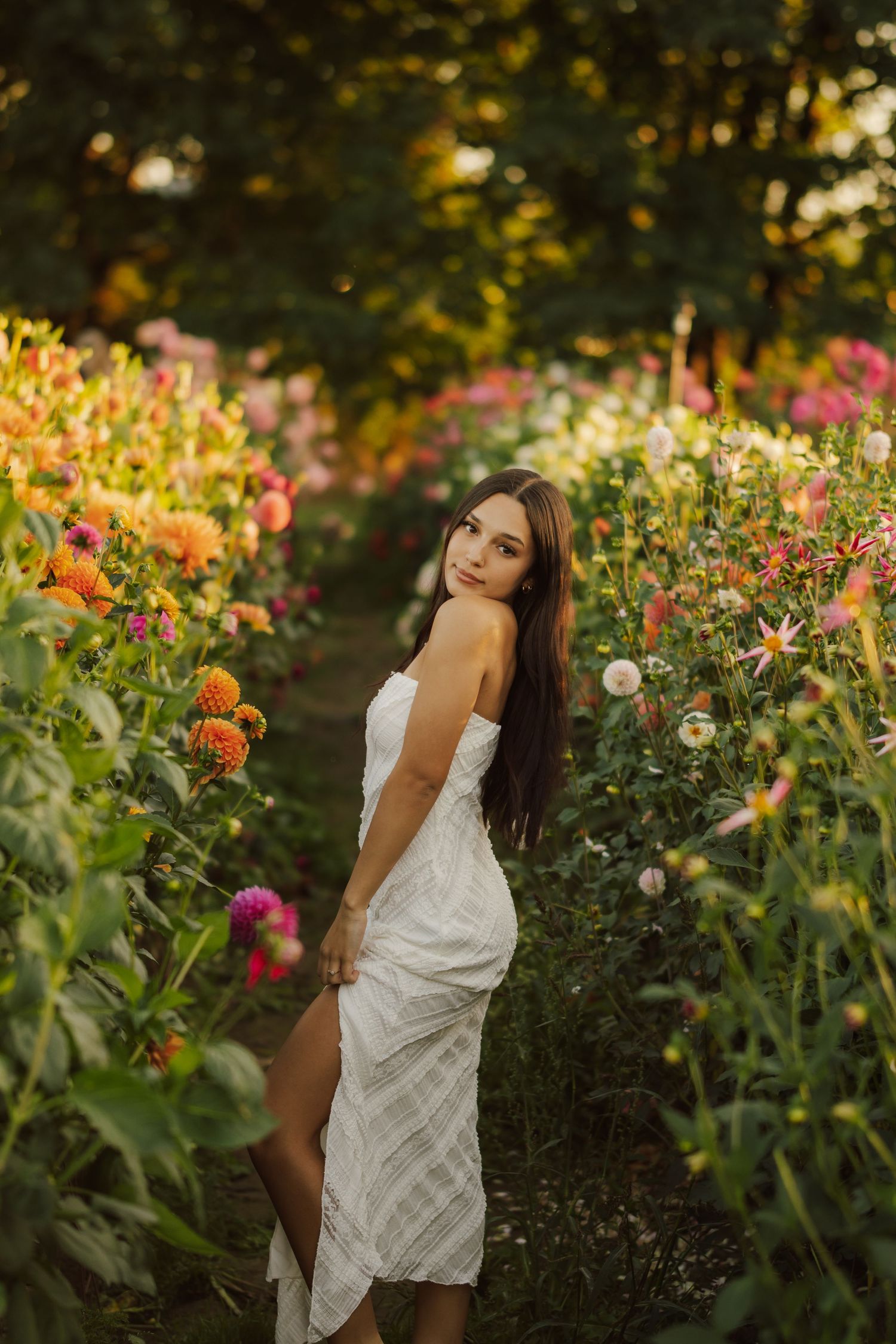 Woman with long dark hair in a white striped sleeveless dress posing on a path between blooming orange and pink dahlias in a flower garden.