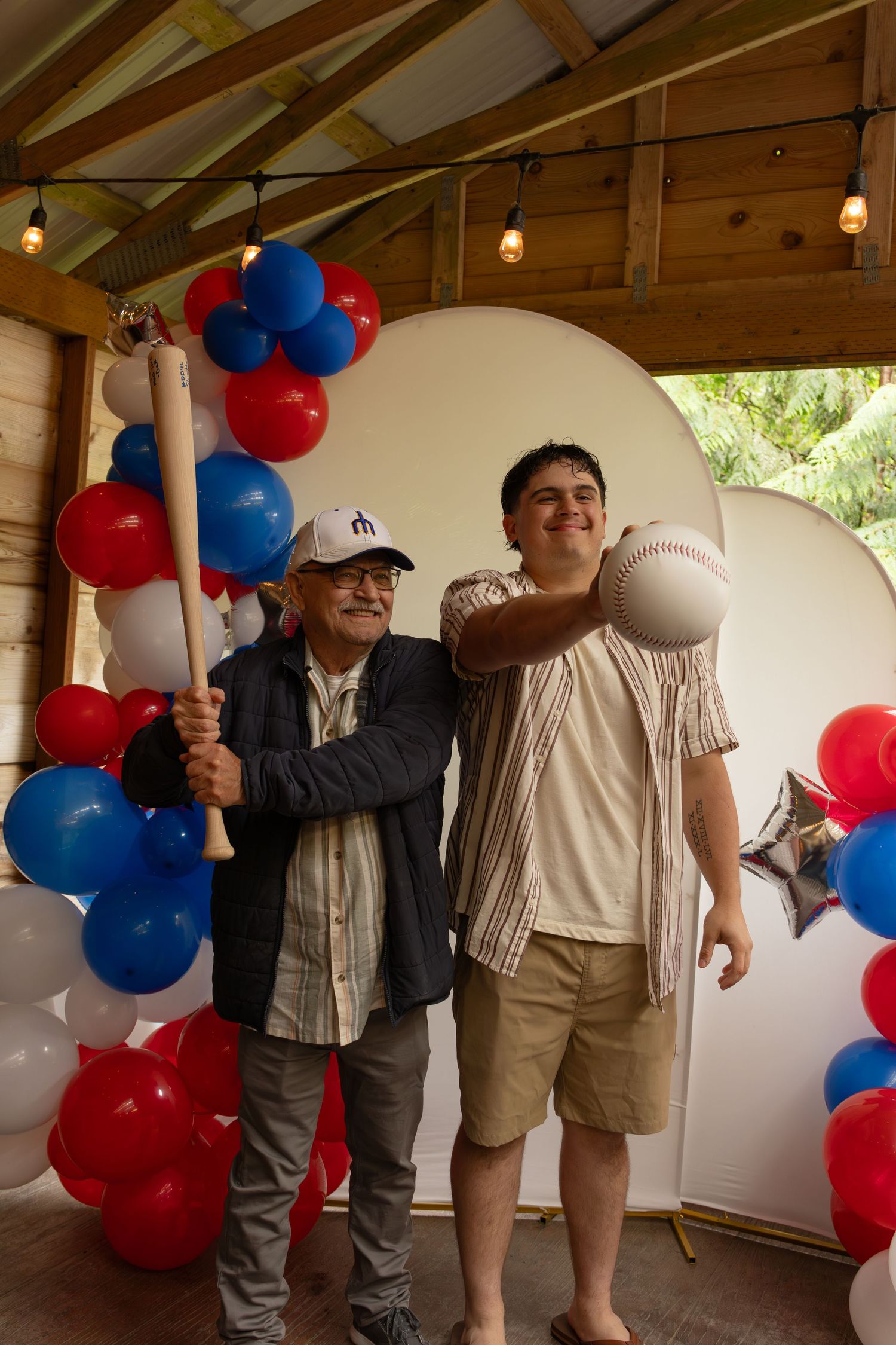 Two men posing with baseball bat and ball near red, white, and blue balloon decorations.
