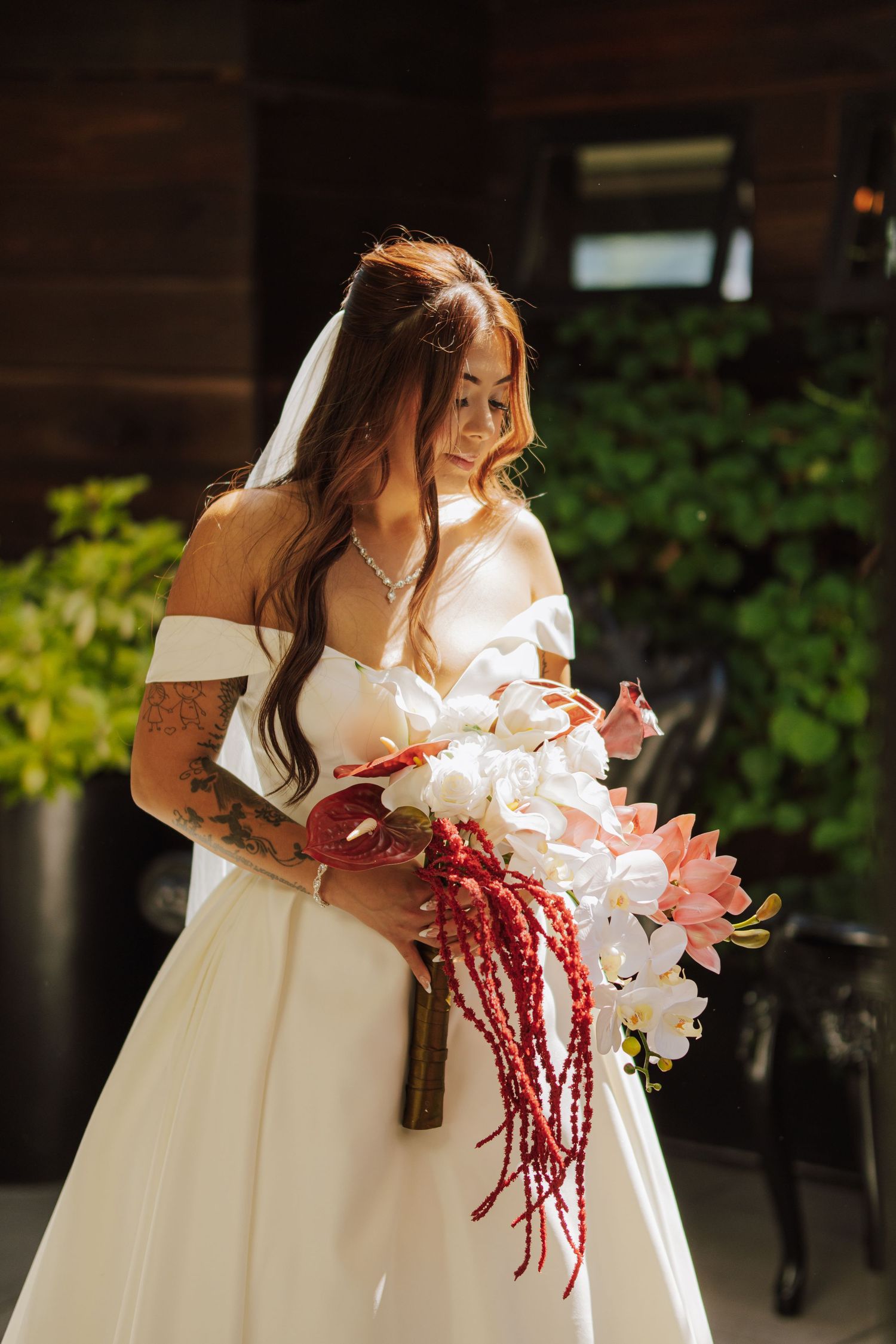 Bride in off-shoulder white gown holding cascading bouquet of white orchids and red amaranthus.