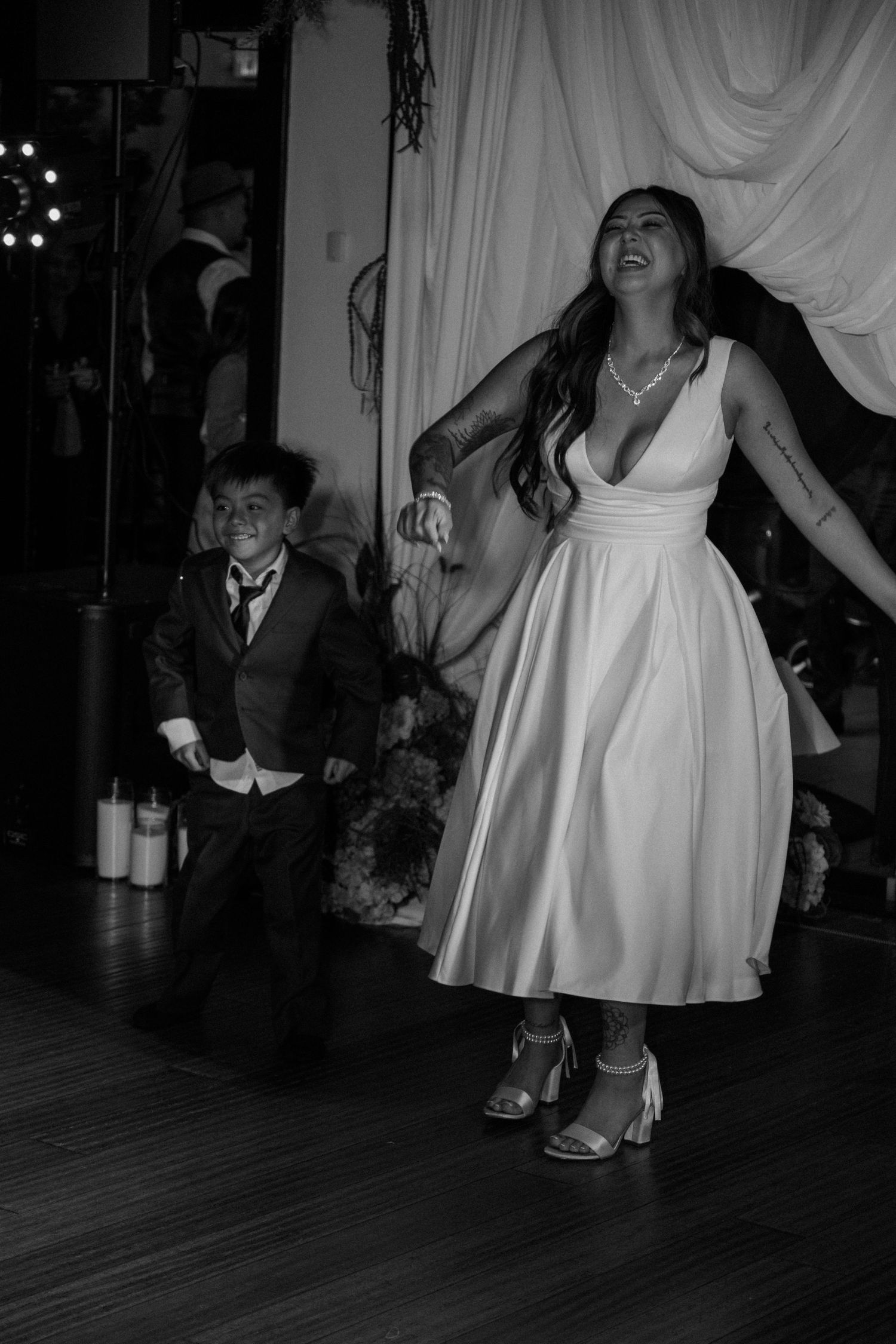 Bride dances energetically with young boy in suit on wedding reception dance floor.