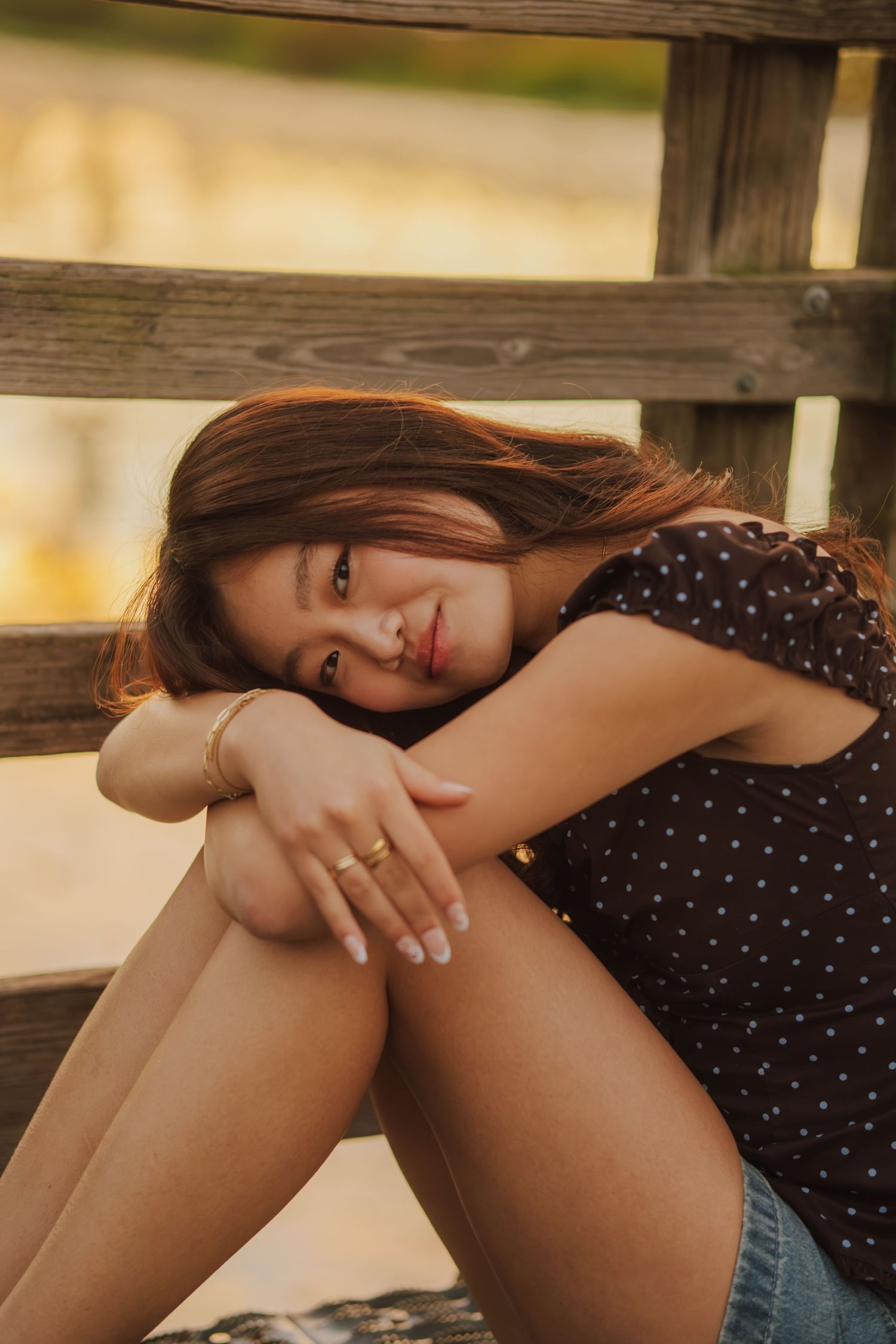 Young woman with long brown hair rests her head on her arms while sitting by a weathered wooden railing in warm golden light.
