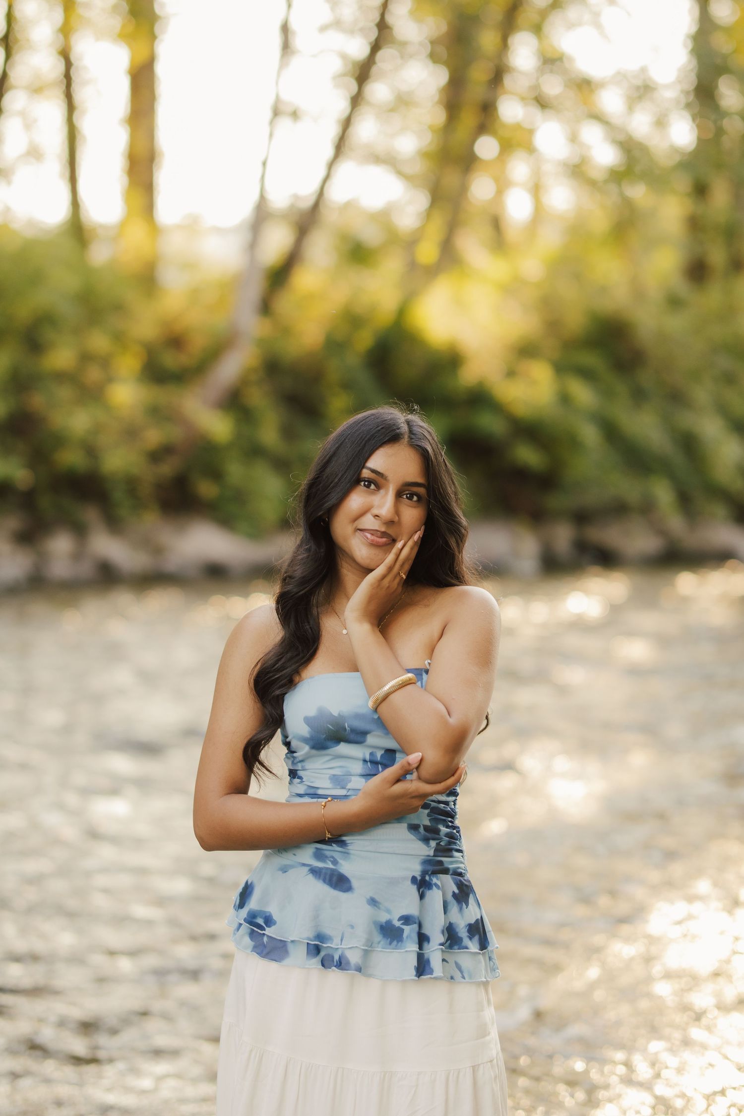 Young woman with long dark hair poses in a blue floral strapless top and white skirt beside a sunlit river with golden trees behind her.