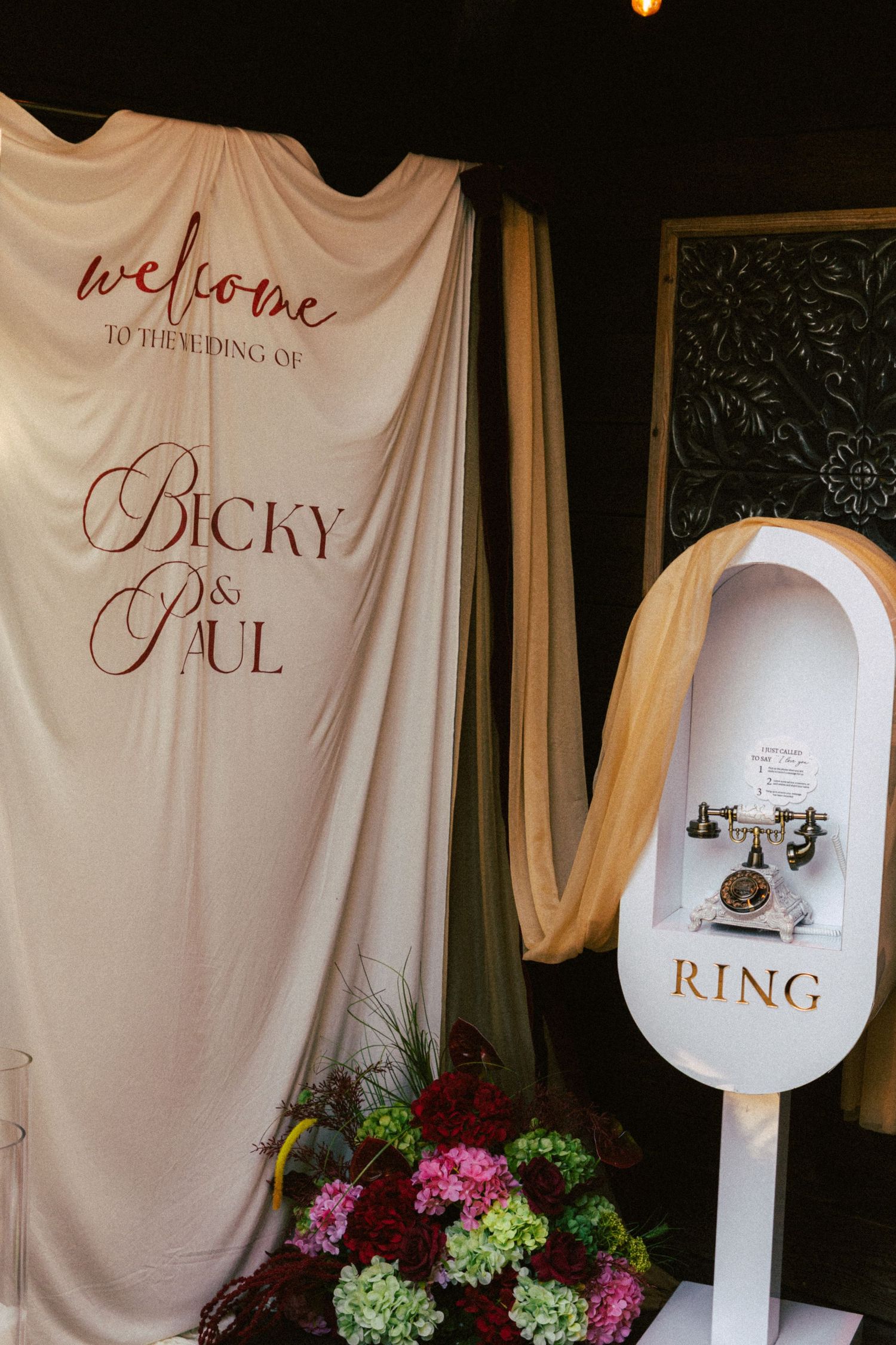 Welcome sign reading Bicky and Paul displayed at wedding entrance with floral arrangements and vintage phone booth.