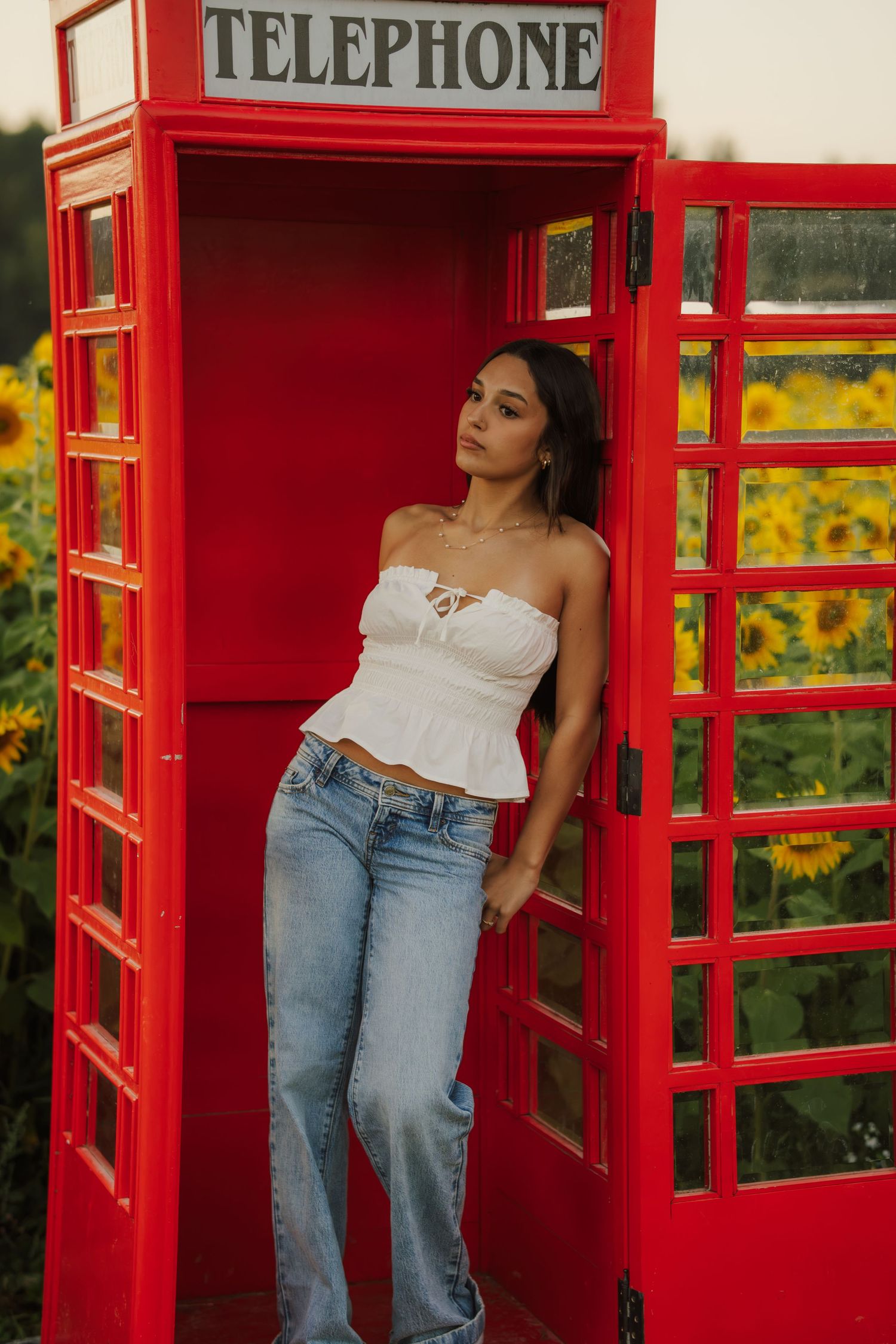 Woman in a white off-shoulder top and light-wash jeans leans against a red British telephone box beside a blooming sunflower field.