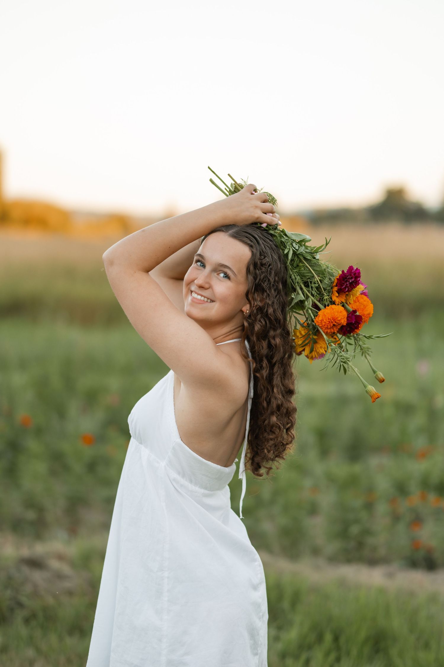 Smiling young woman in white dress holding colorful wildflower bouquet overhead in a sunlit green field at golden hour.