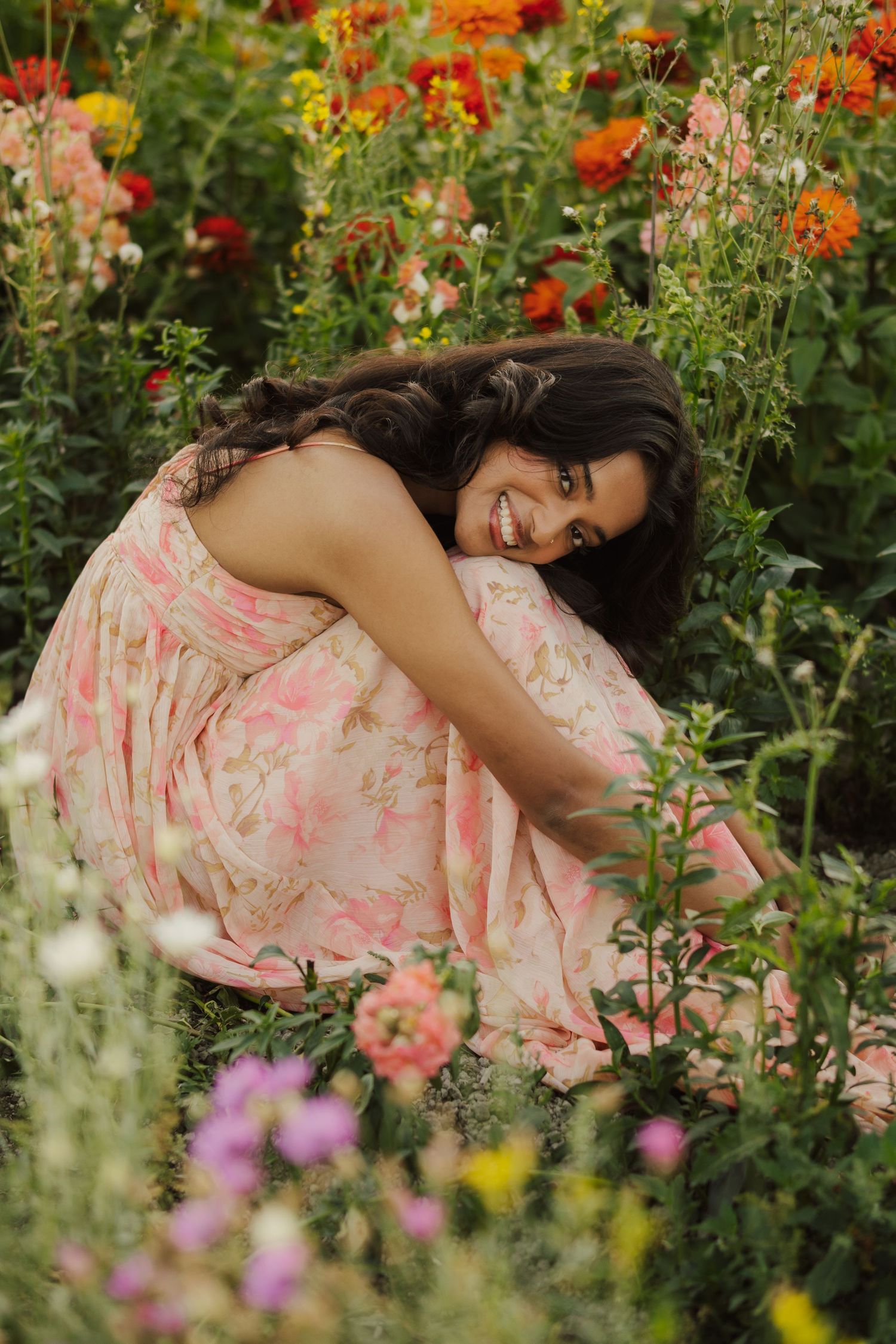 Smiling woman with long dark hair in a pink floral dress crouching among colorful orange, pink and purple garden flowers.