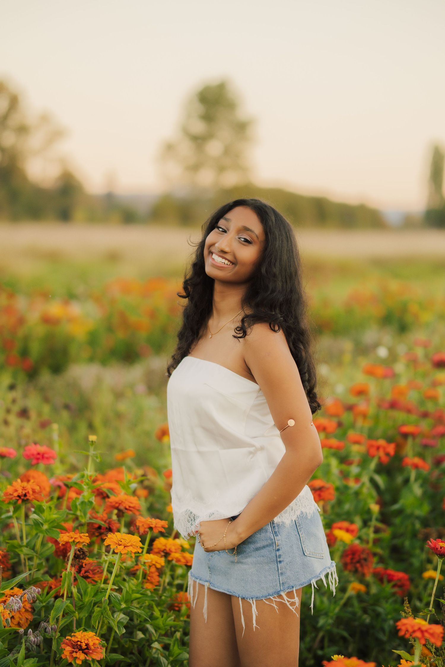 Smiling young woman with long dark curly hair in a white top and denim shorts standing among orange zinnia flowers in a field at sunset.