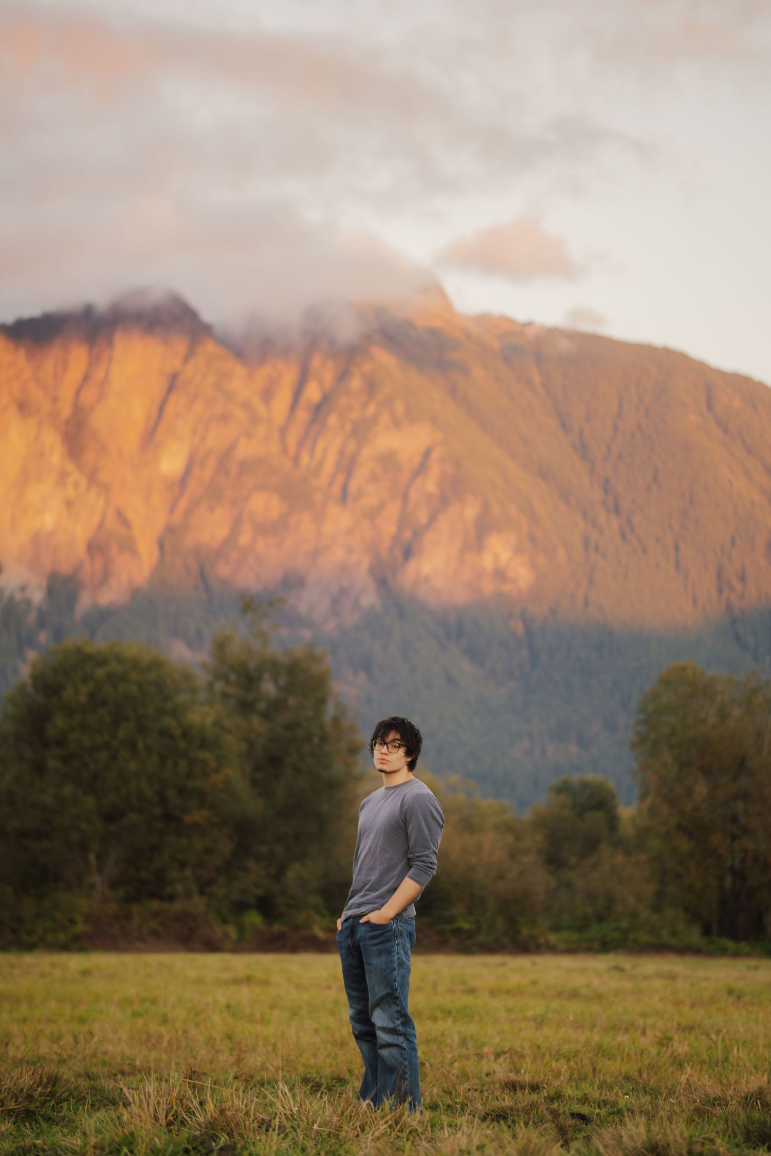 Man in a gray sweater and jeans stands in a grassy field at sunset with a glowing orange mountain and misty clouds behind him.