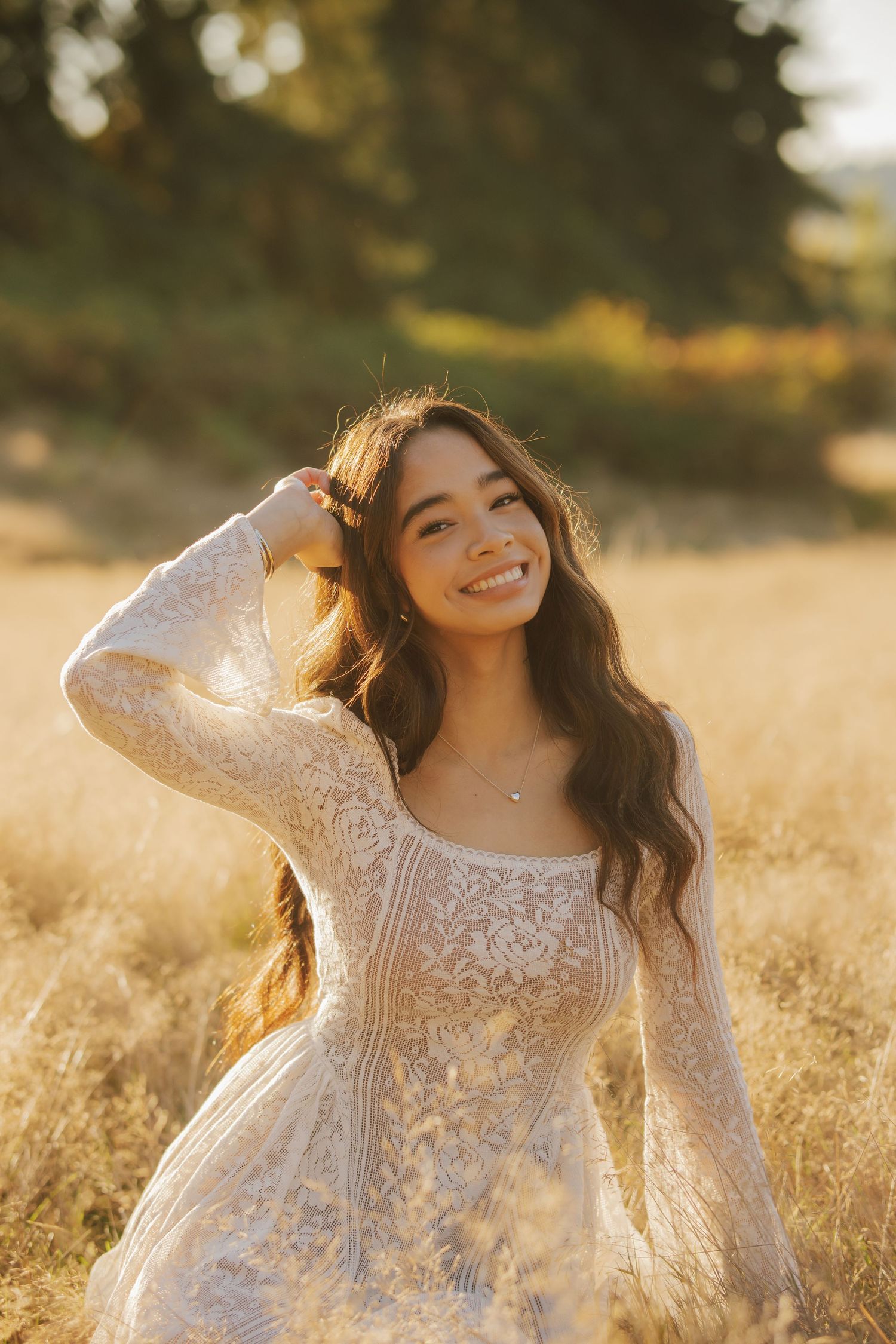 Smiling woman with long wavy hair in a white lace dress sits in a golden meadow, one hand in her hair, bathed in warm sunset light.