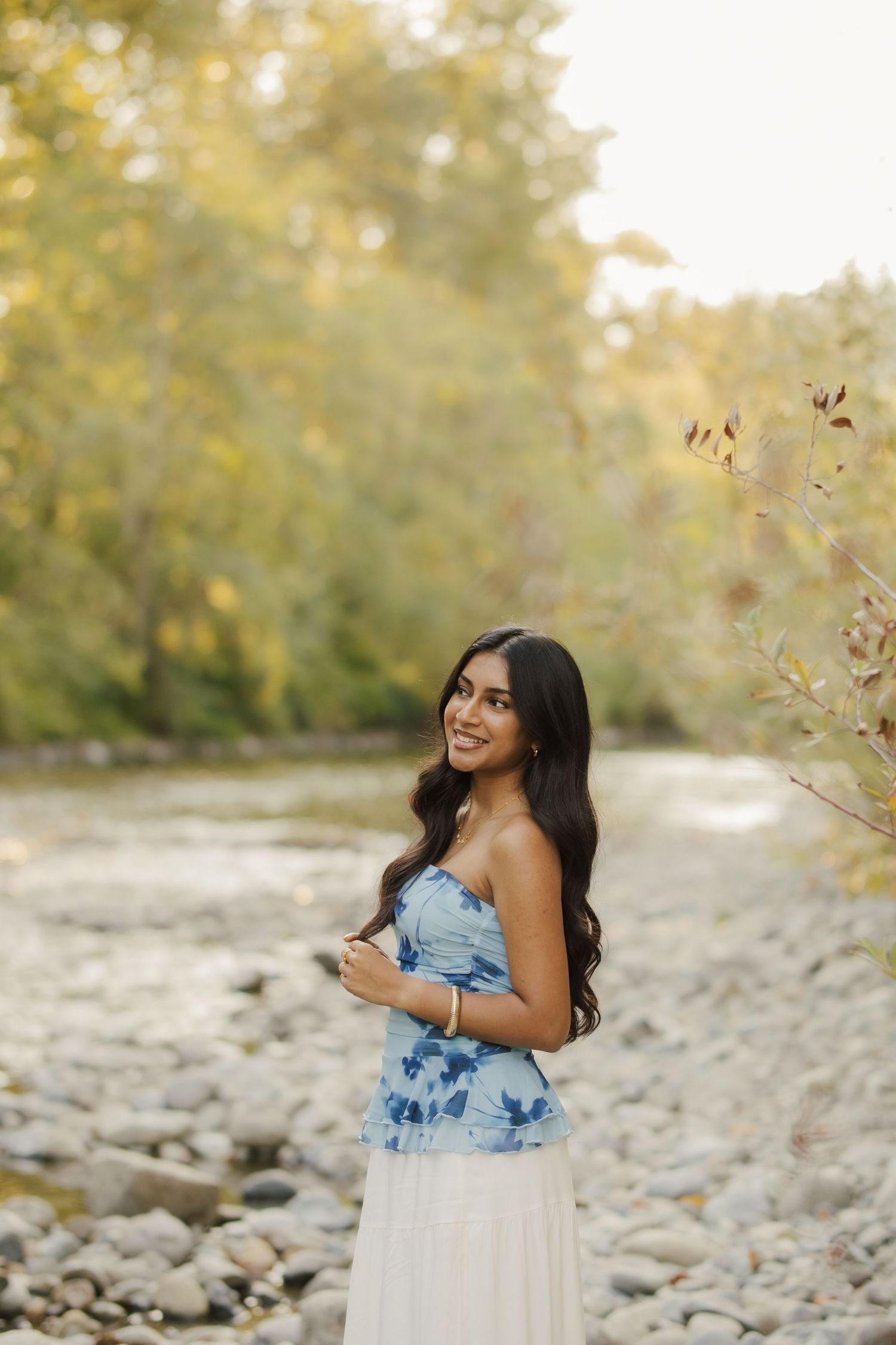 Young woman in blue floral top and white skirt smiling by a rocky riverbank surrounded by autumn trees.