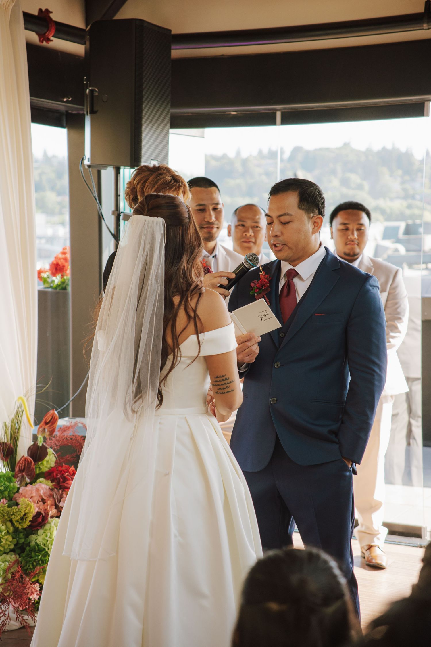 Groom in navy suit reads vows to bride at altar surrounded by groomsmen and colorful floral decor.