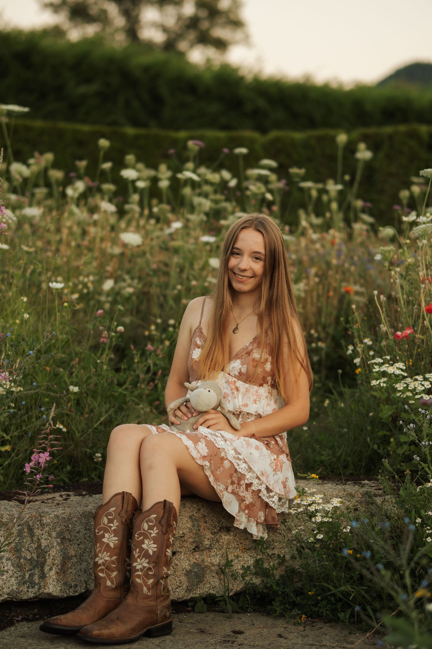 Smiling teenage girl in floral dress and brown cowboy boots sitting on stone wall in wildflower garden.