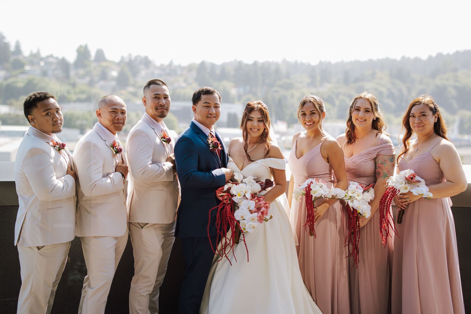 Wedding party poses outdoors, bridesmaids in blush pink gowns and groomsmen in tan suits with bride and groom centered.