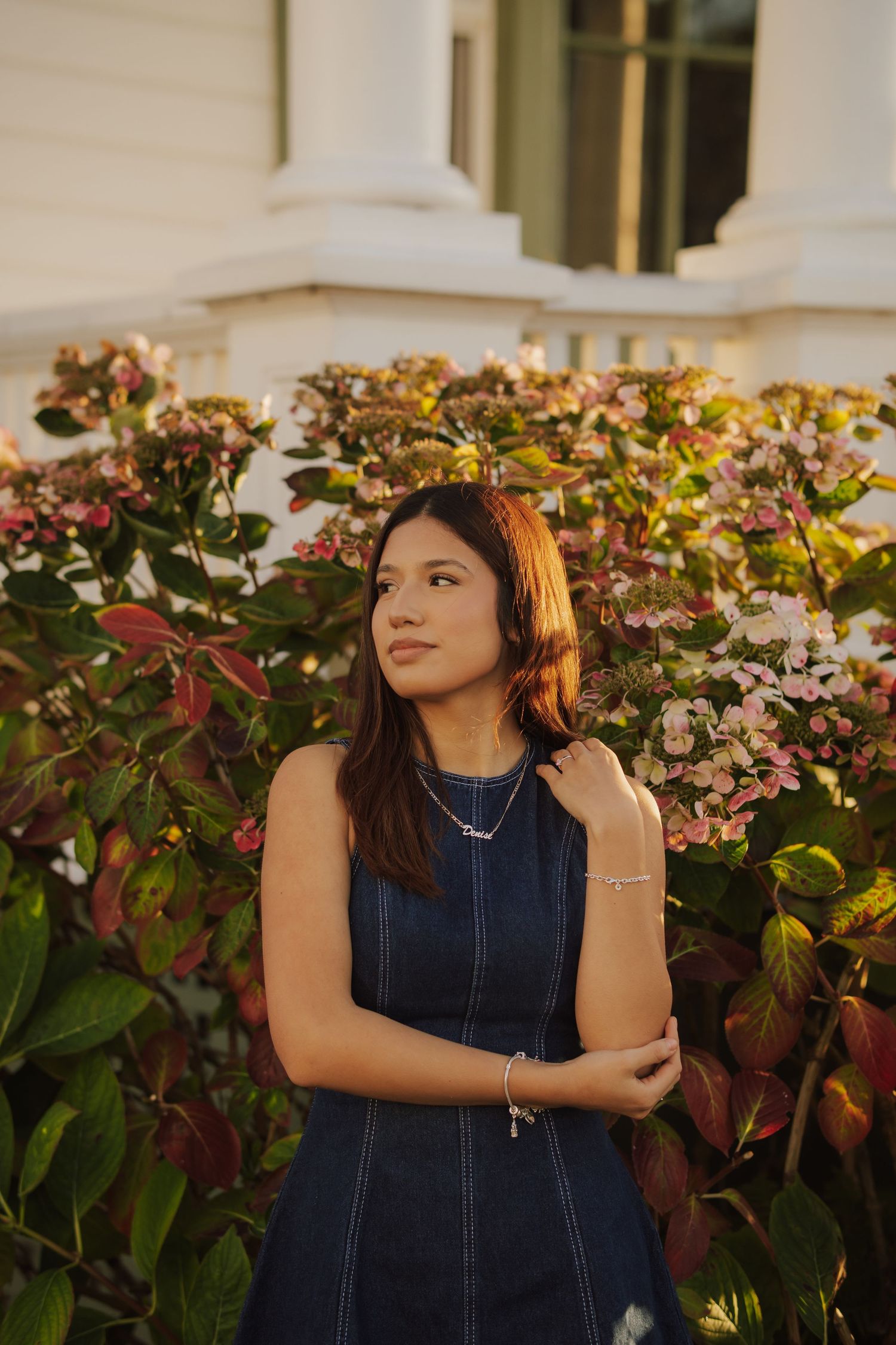 Young woman with long dark hair in a denim sleeveless dress poses in golden light beside blooming pink hydrangeas outside a white building.
