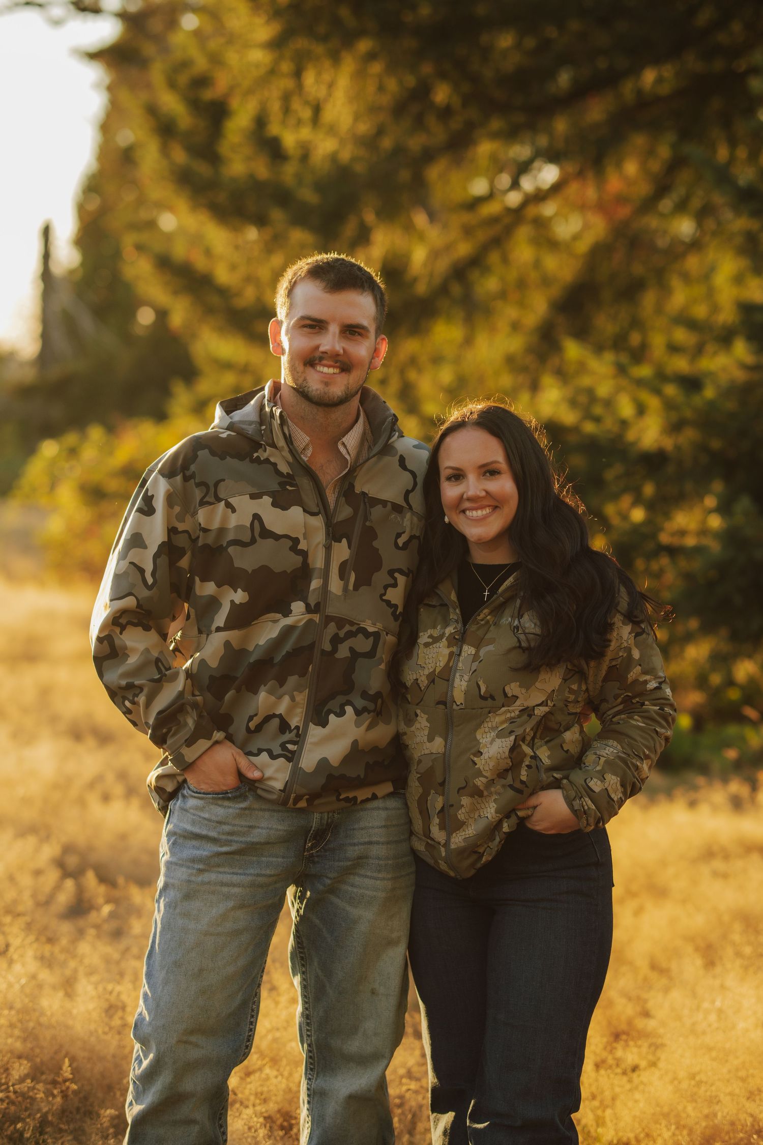 Smiling man and woman in matching camouflage jackets and jeans pose together in a golden grassy field with sunlit trees behind them.