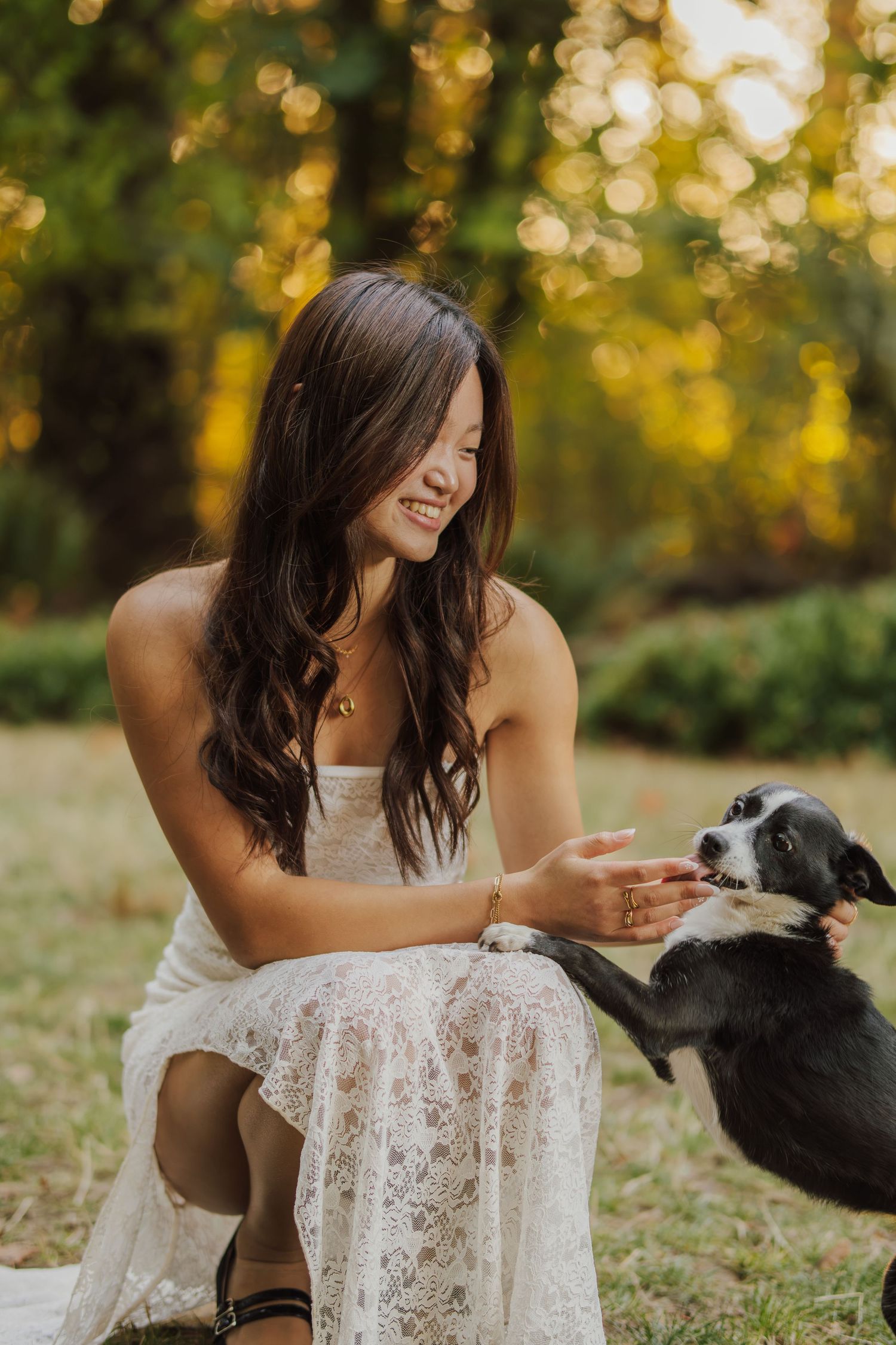 Smiling woman in a white lace dress crouches on a grassy lawn as a black and white dog raises its paw to her hand in golden sunlight.