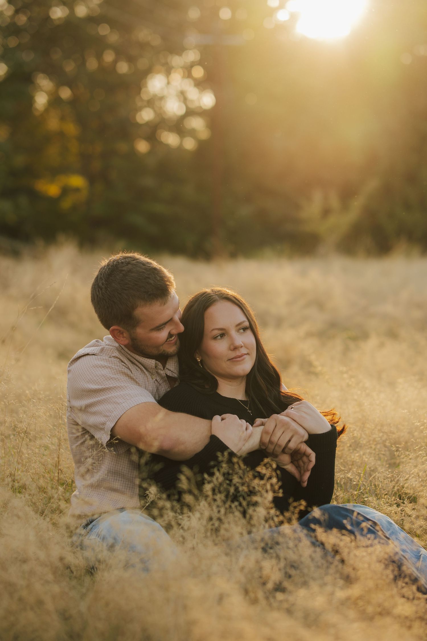 Couple sitting close together in a golden grassy field at sunset, the man embracing the woman from behind in warm, glowing light.