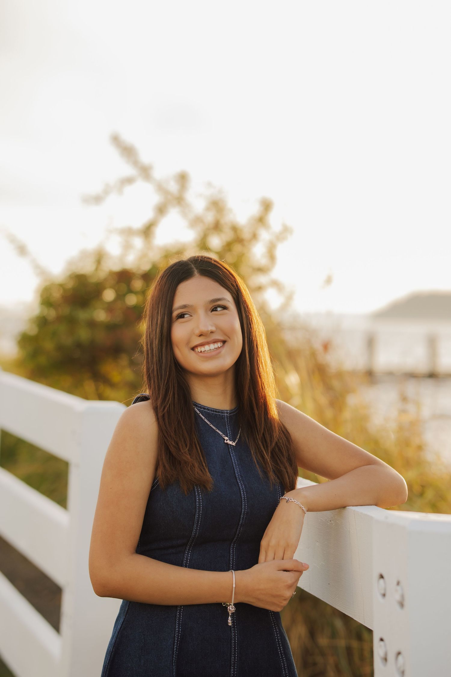 Smiling young woman with long brown hair in a denim dress leans on a white wooden fence at sunset with golden grass and water behind her.