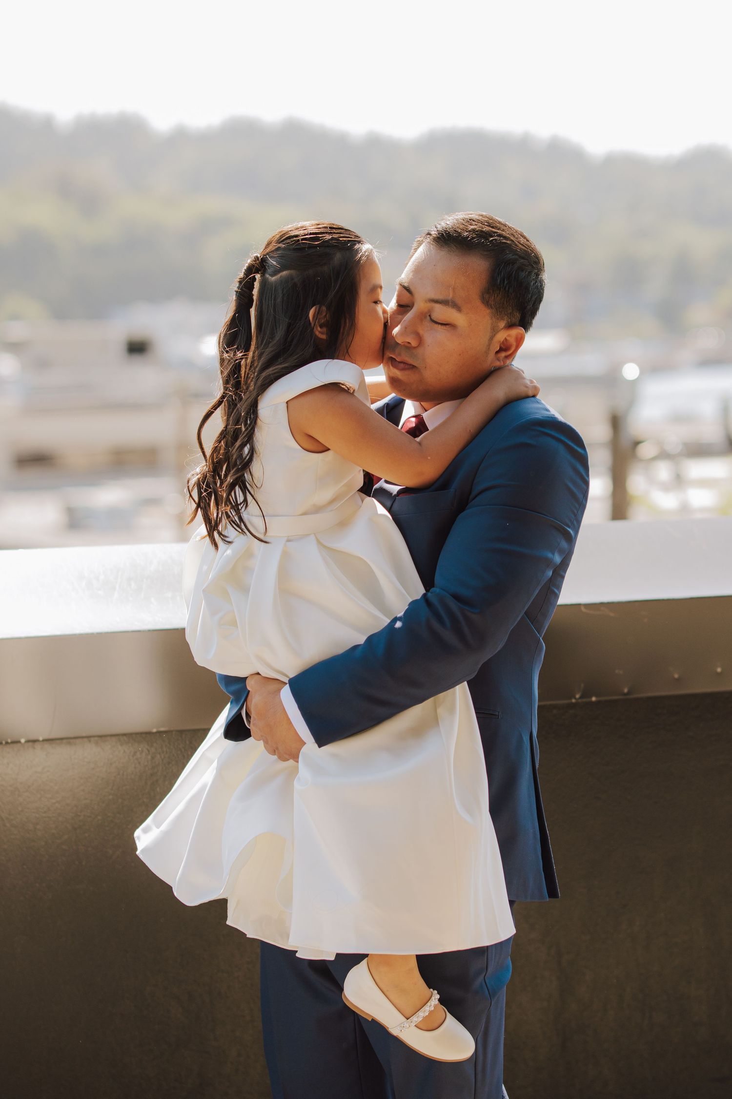 Groom in navy suit tenderly holding a young girl in a white dress on a rooftop.