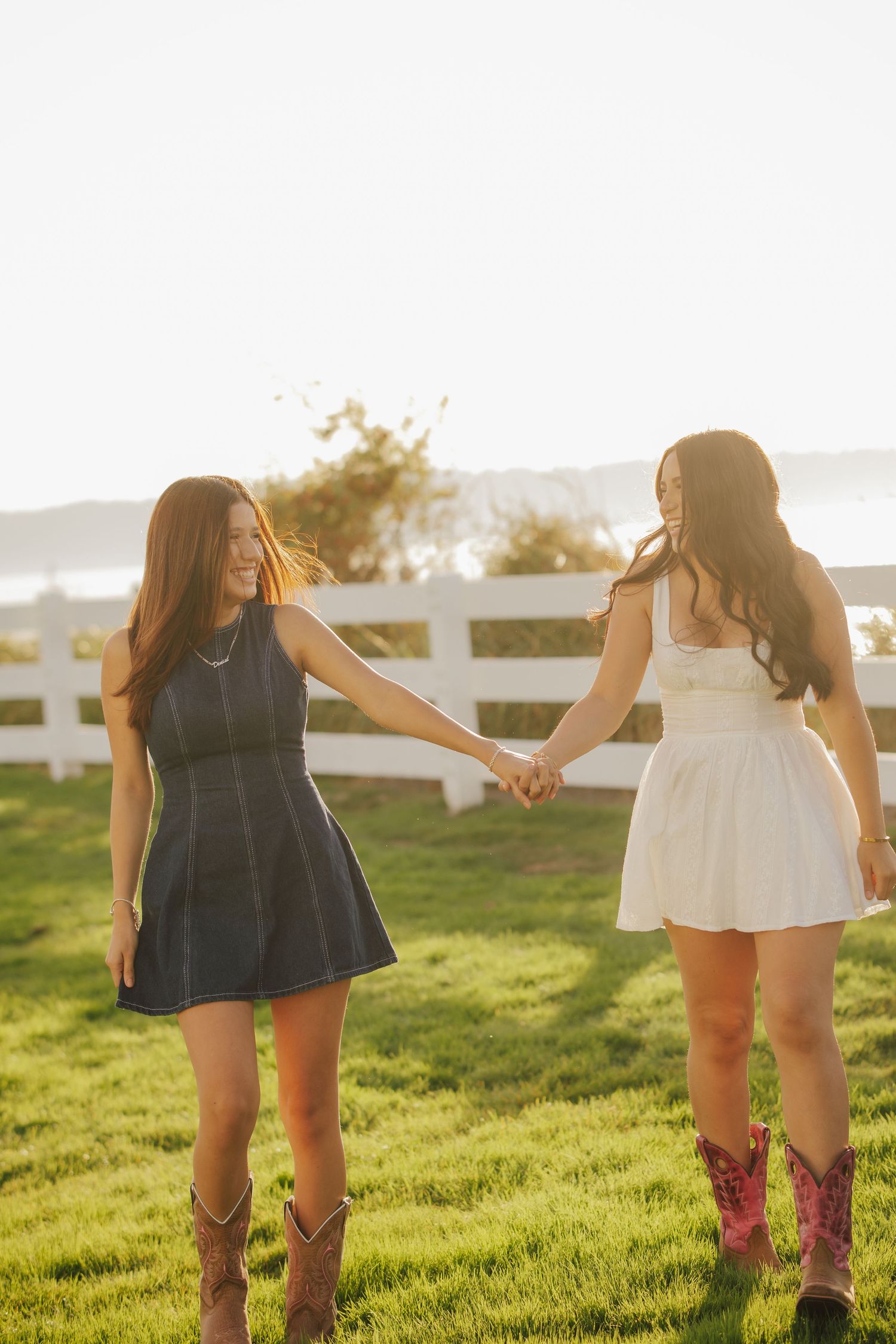 Two young women in dresses and cowboy boots hold hands and walk across a sunny grassy field near a white fence at golden hour.