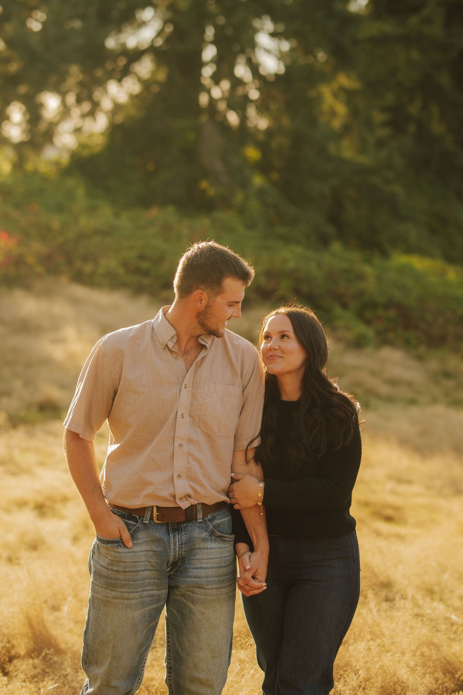 Smiling couple holding hands and gazing at each other while walking through a golden grassy field in warm evening sunlight.