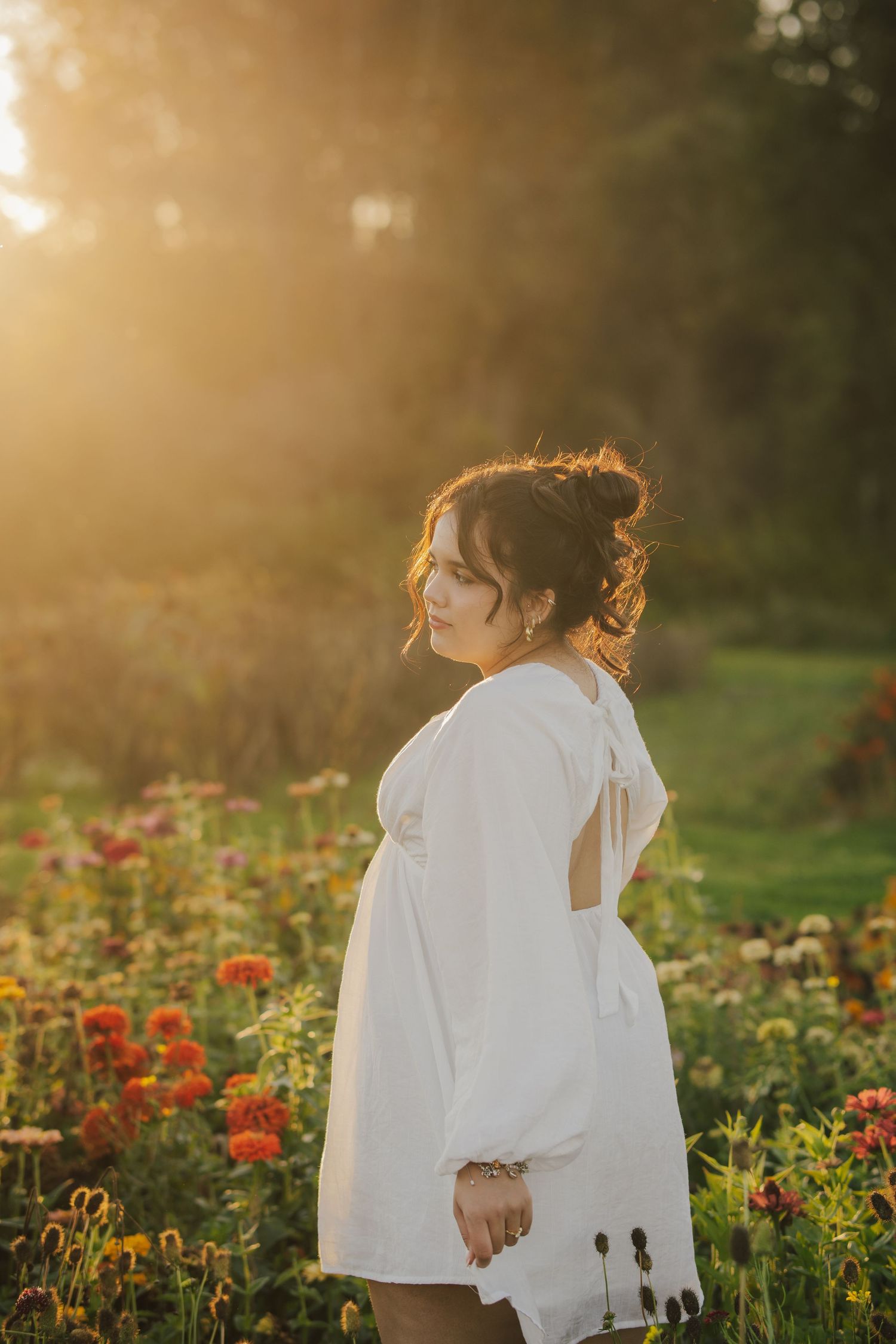 Woman in a white open-back dress with an updo stands in profile among orange and red zinnias in a field, lit by warm golden sunlight.