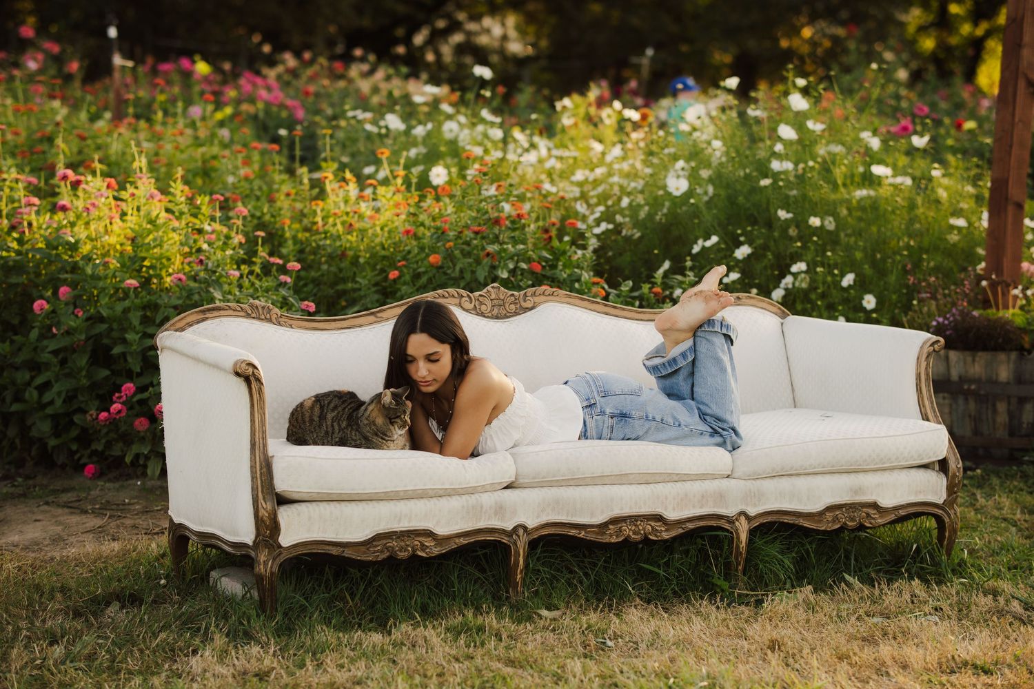 Woman with long dark hair relaxes on a cream vintage sofa with a cat in a sunny garden surrounded by colorful wildflowers.