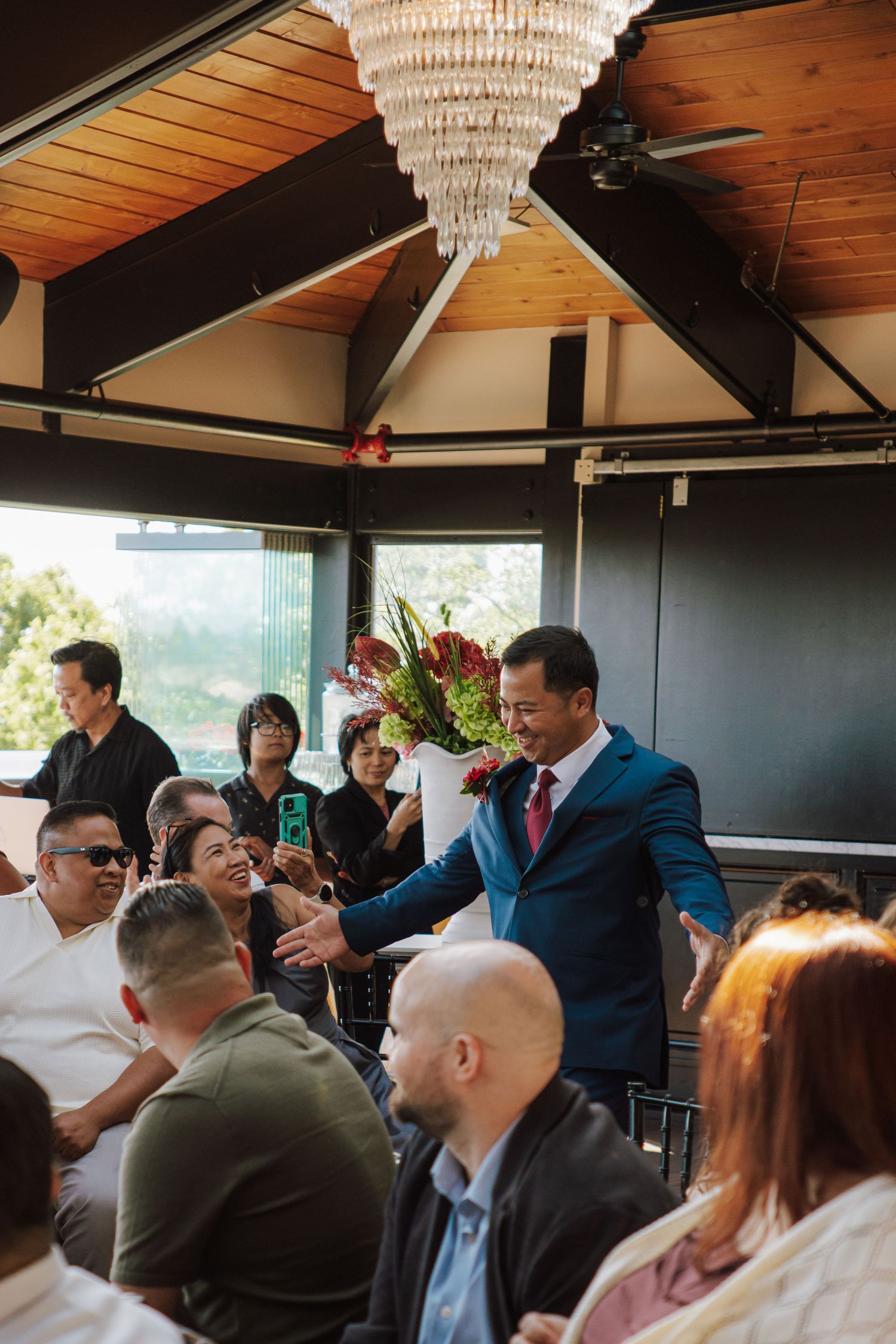 Groom in navy blue suit greets seated wedding guests inside elegant venue with chandelier overhead.