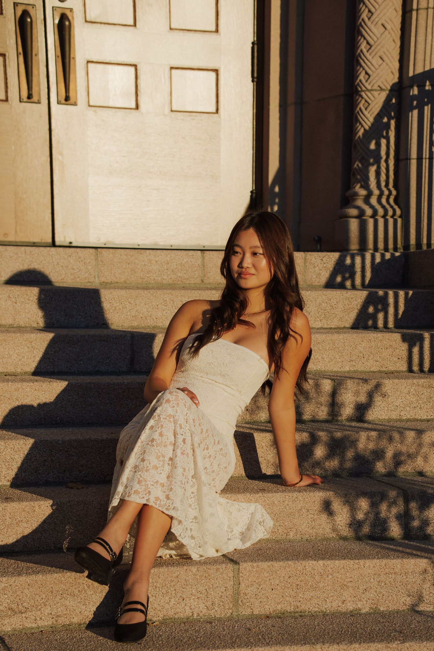 Woman in a white strapless top and floral skirt sits on sunlit stone steps in golden light, wearing black Mary Jane shoes.
